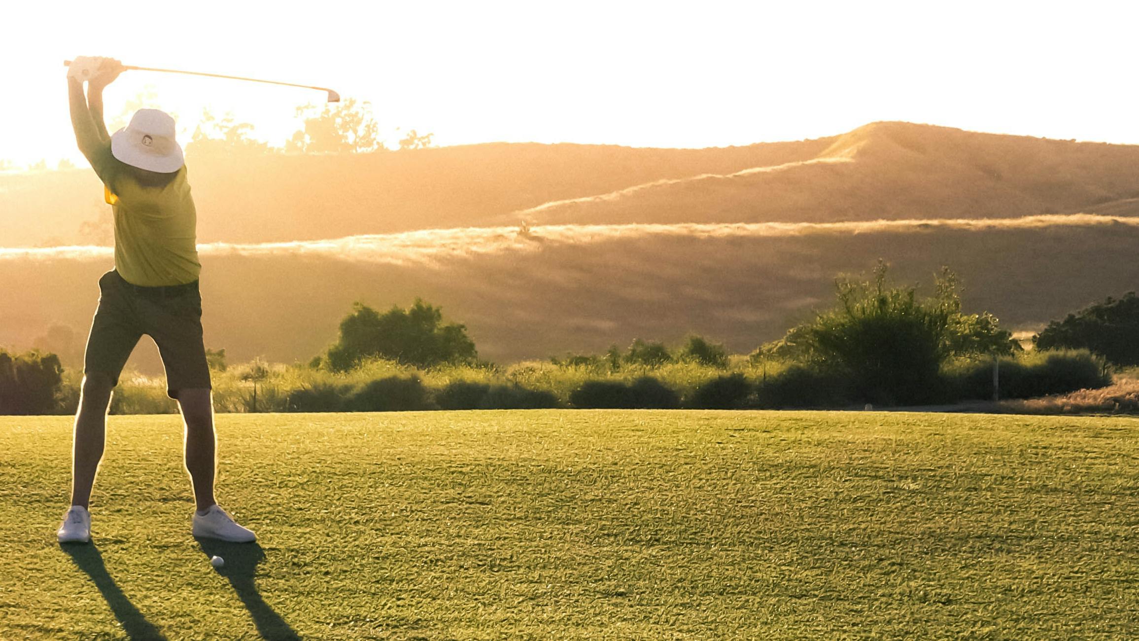 A man swings at a golf ball while the sun sets behind him.