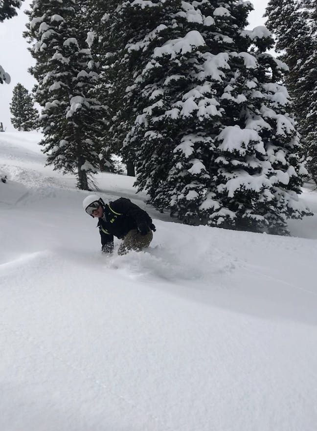 A snowboarder takes a turn down a snowy trail. He is wearing a backpack and the Smith Holt helmet in white.