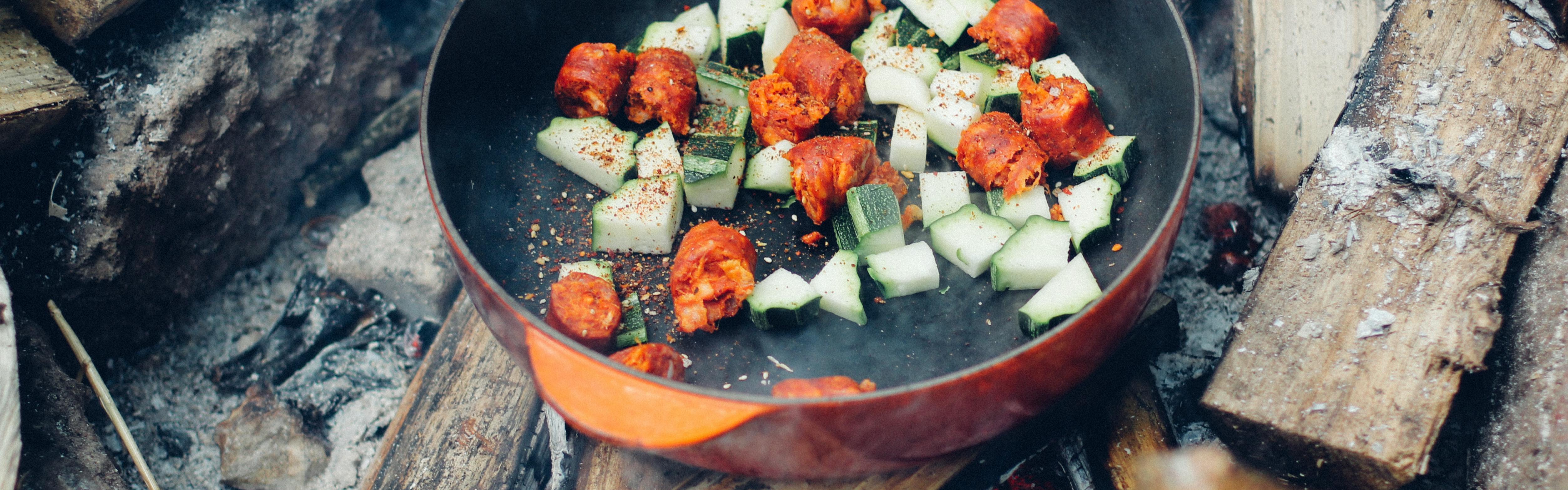 Cut-up zucchini and sausage sit in a pan over a campfire. 