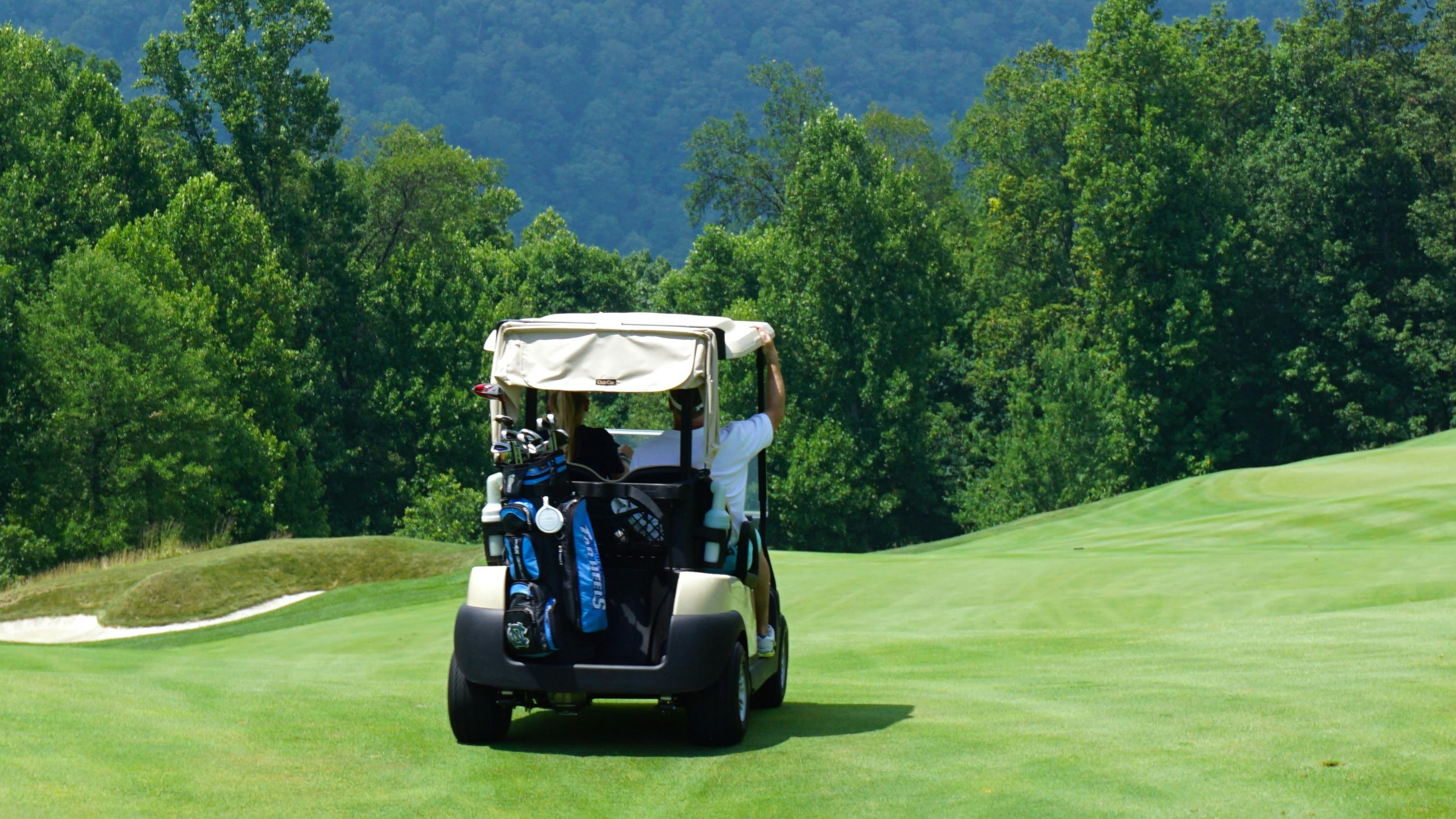 Two people drive away on a golf cart towards a tree-lined golf course