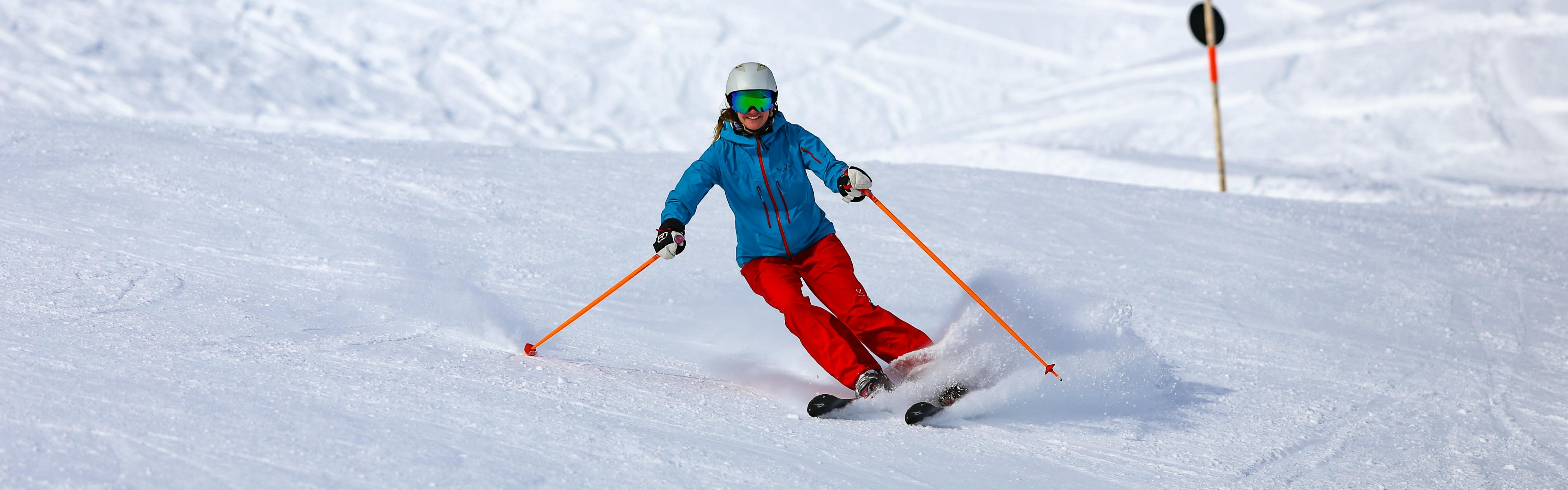 A woman skis down a slope holding two orange poles and wearing a helmet, goggles, red pants, and a blue jacket. 