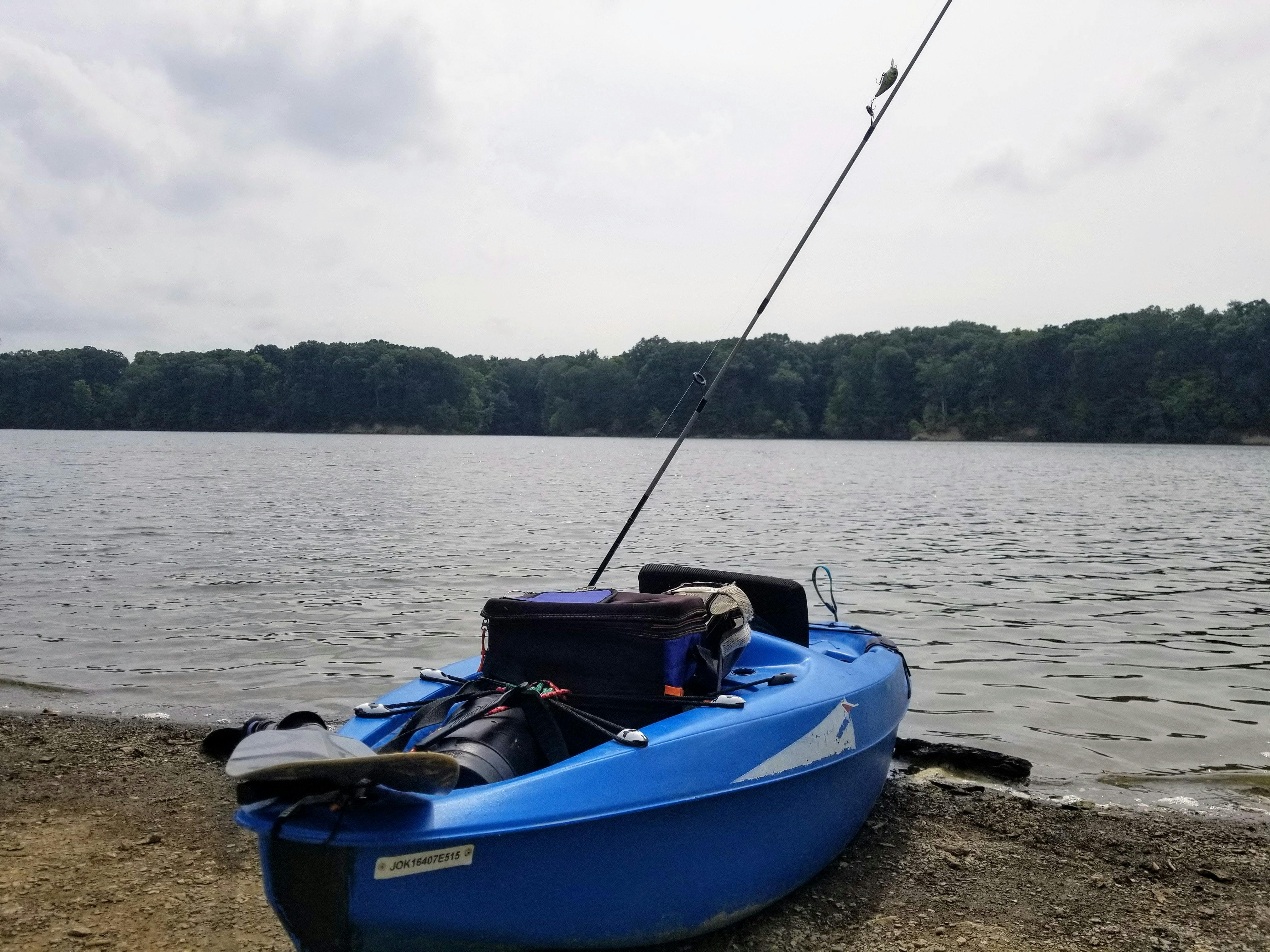 A kayak sits on the shore of a lake with a fishing rod sticking up out of it. A PFD and other miscellaneous gear are sitting inside. 