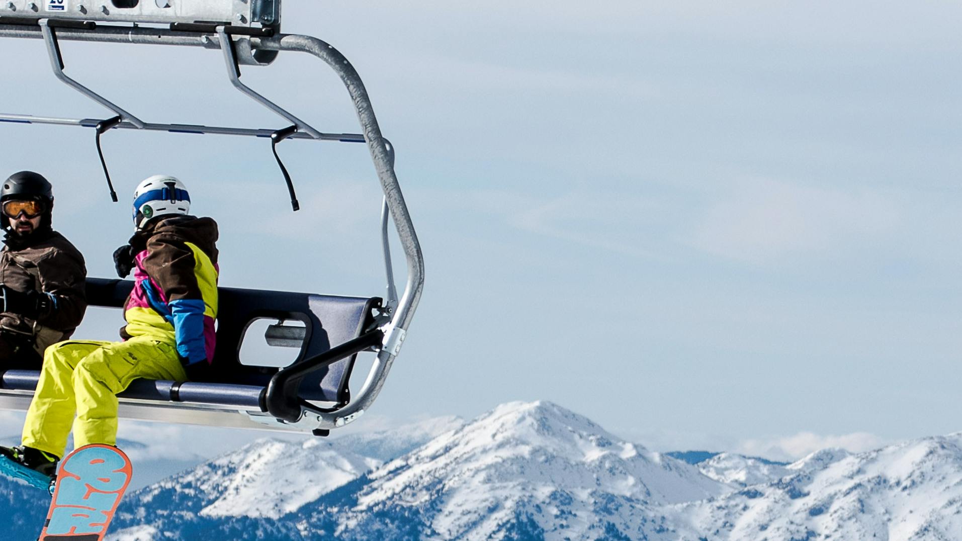 Two snowboarders sitting on a chairlift with mountains in the background.