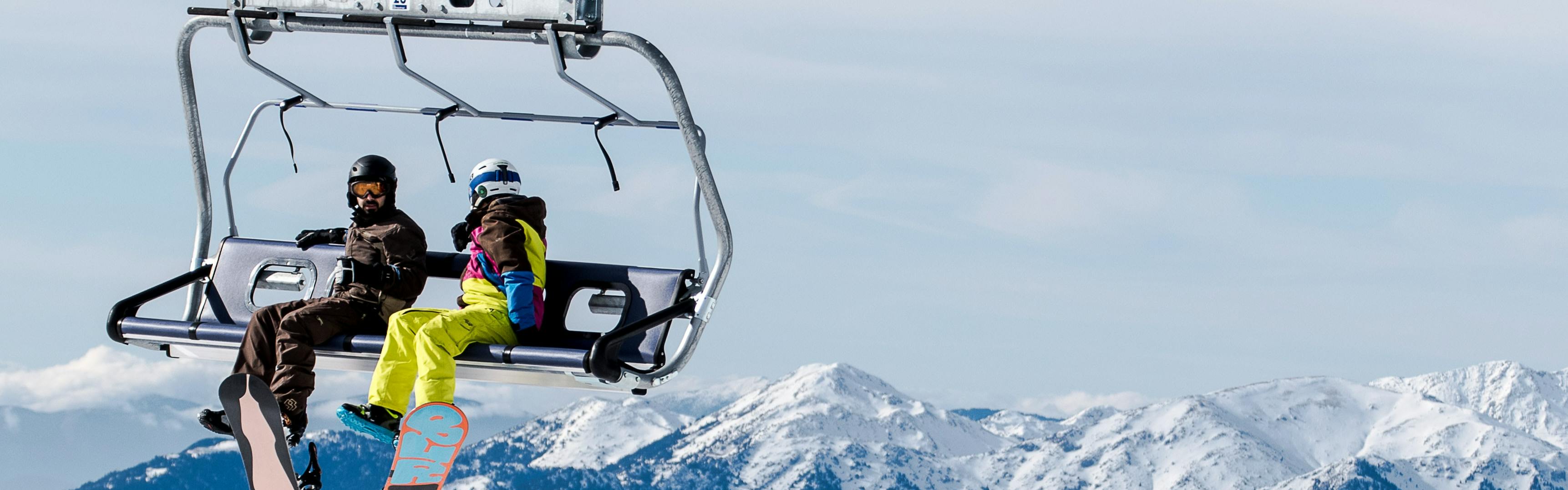 Two snowboarders sitting on a chairlift with mountains in the background.