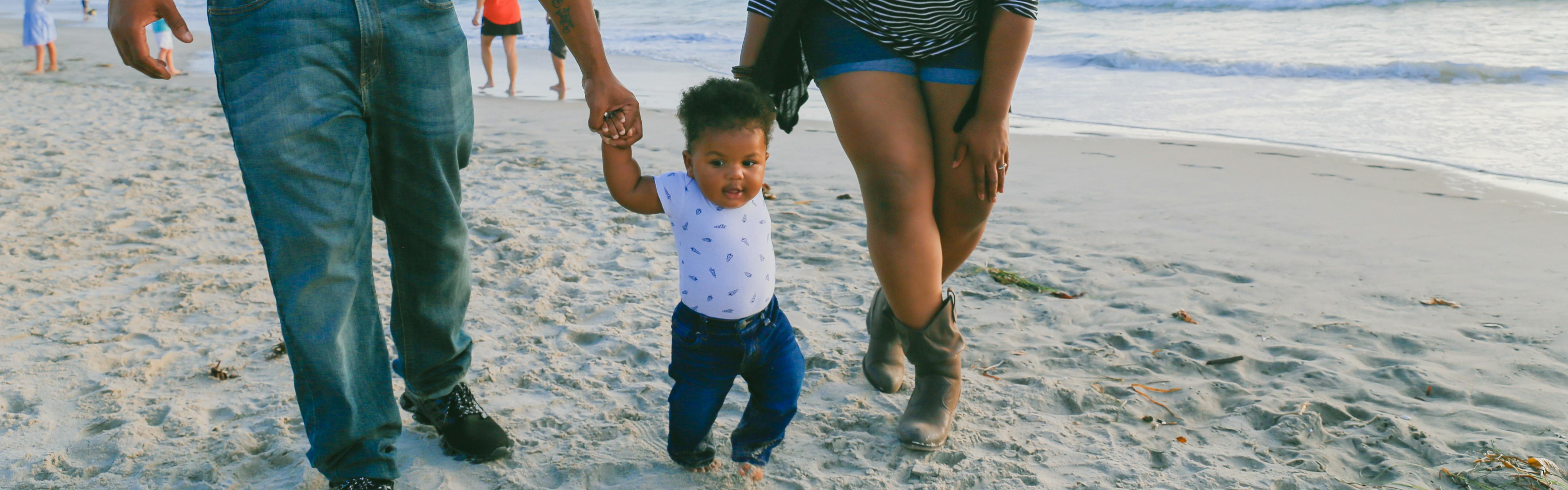 A baby walking in the sand. Her hands are each being held by her parents as they walk next to her. 