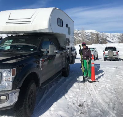 A woman standing with her skis in a ski parking lot next to a truck with a camper.