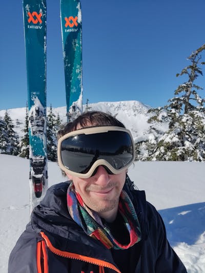 Selfie of a man with goggles on standing in a snowfield with the Volkl Blaze 106 skis behind him.