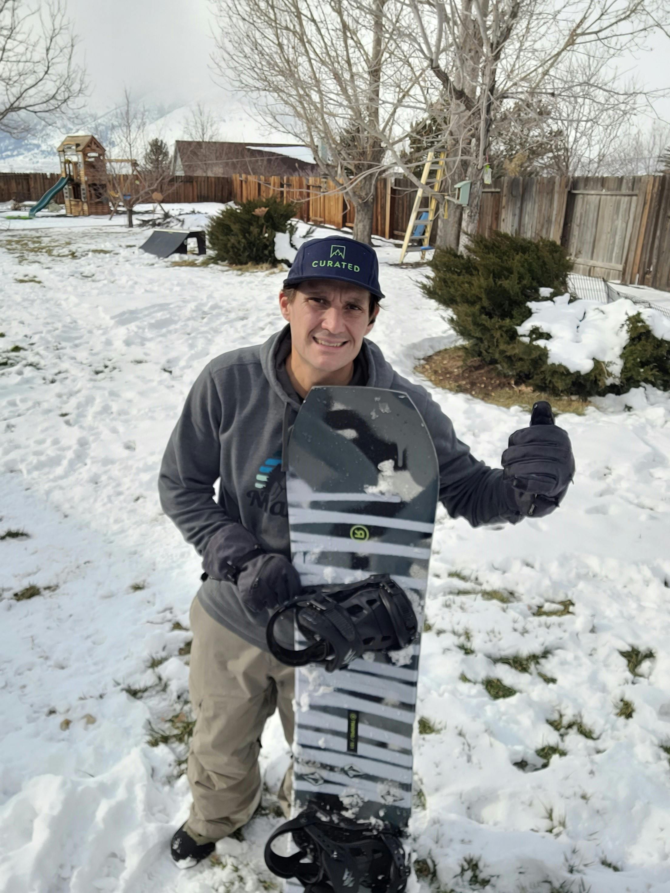 A man standing in snow with a thumbs up. He is holding a Ride Twinpig Snowboard.