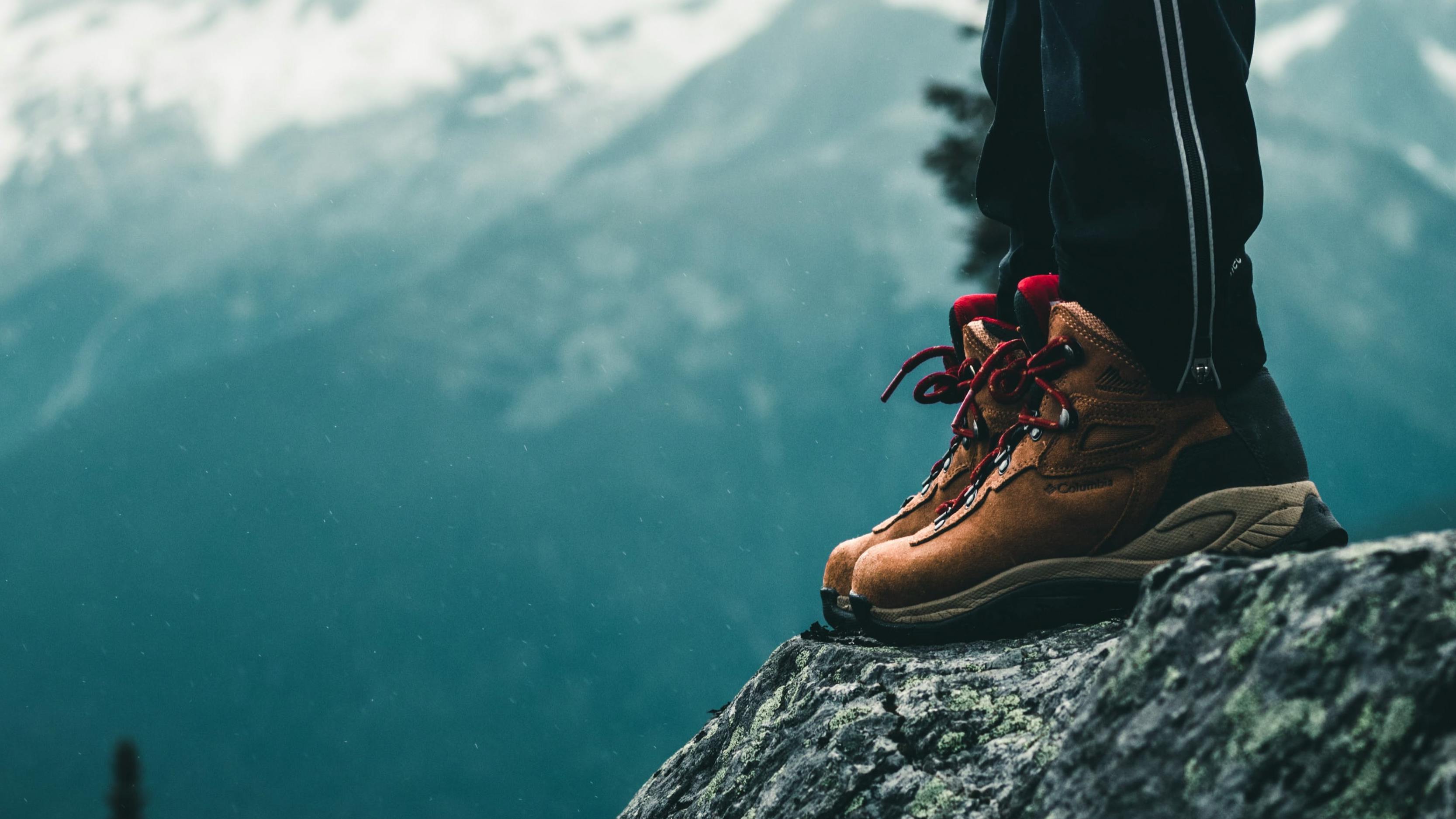 A close up of a pair of hiking boots as a hiker stands on the edge of a rock. There are trees and snow covered mountains in the background. 