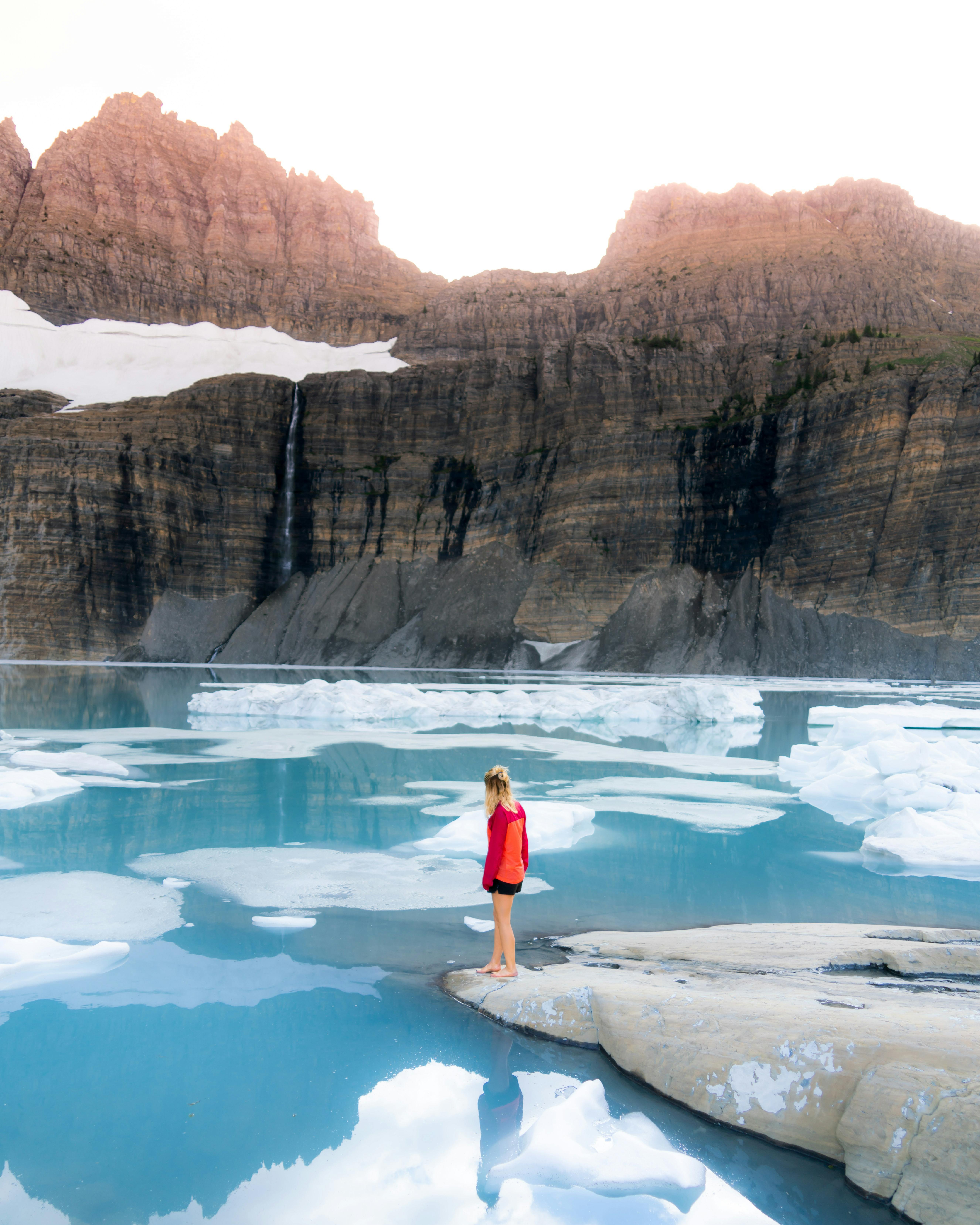 A woman dips her toe in a very blue lake. There is ice floating in the lake. 
