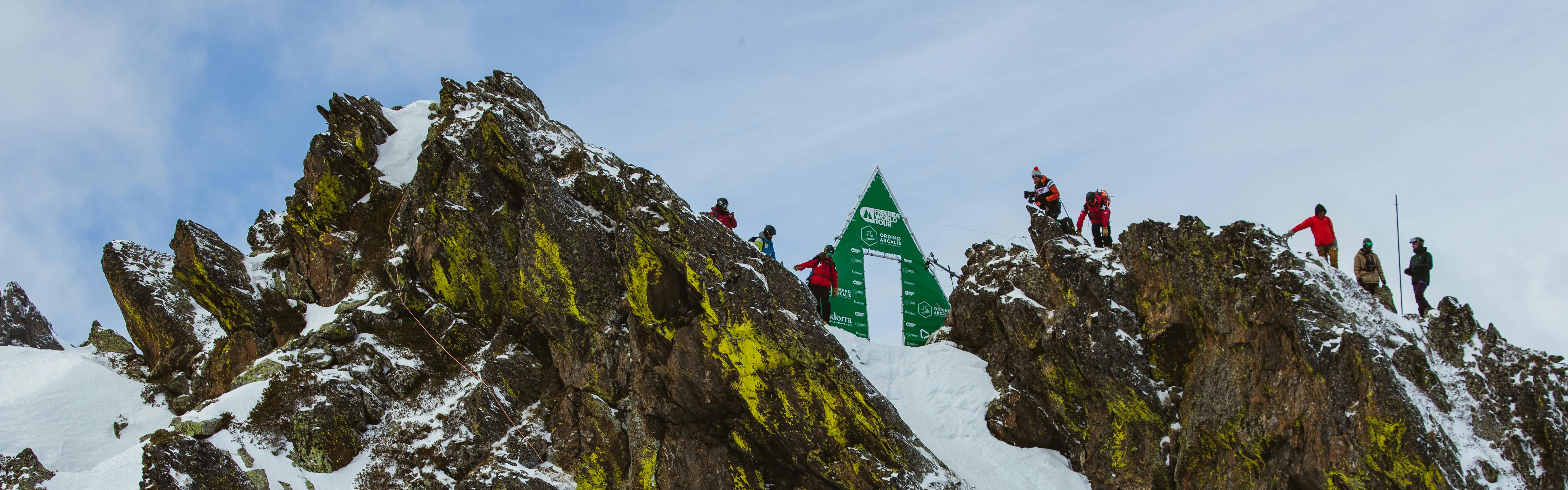 An FWT stand sits on the top of a rocky ridge with skiers clustered around it.