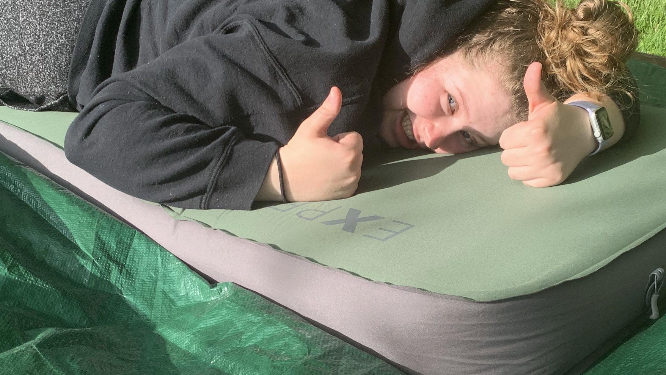 A woman lays on a sleeping bag that is on a tarp giving two thumbs up. 