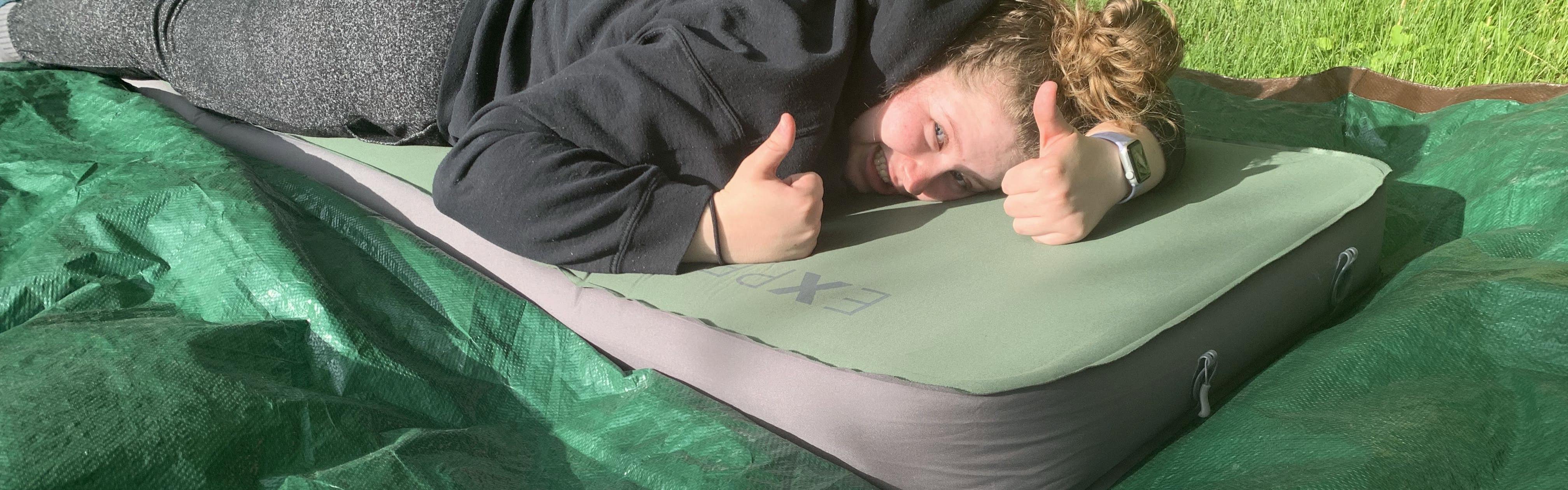 A woman lays on a sleeping bag that is on a tarp giving two thumbs up. 