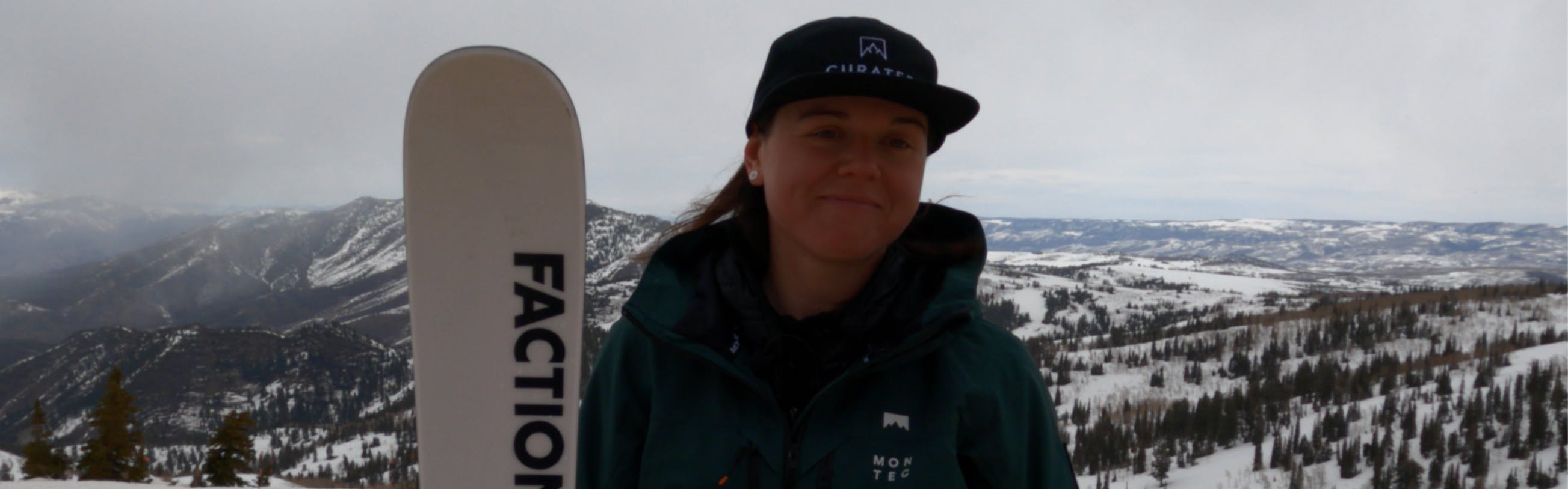A woman standing with the Faction Mana 2X Ski. There is a ski resort and snowy mountains in the background. 