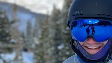 A man wearing the Smith Holt helmet and a pair of goggles smiles at the camera. There are snowy mountains in the background.