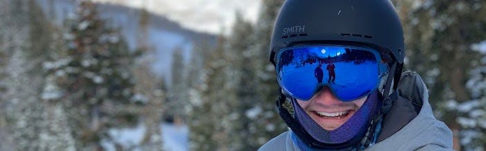 A man wearing the Smith Holt helmet and a pair of goggles smiles at the camera. There are snowy mountains in the background.