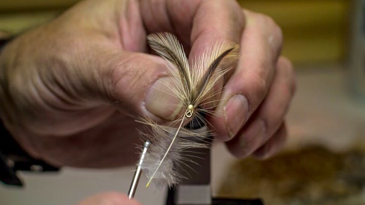 The hands of an older man work on flying a tie with feathers on it.