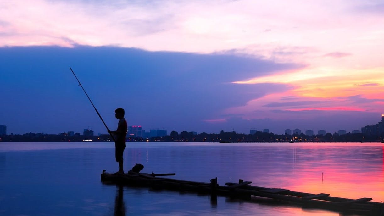 A fisherman standing on a dock with his fishing rod as the sun goes down. 