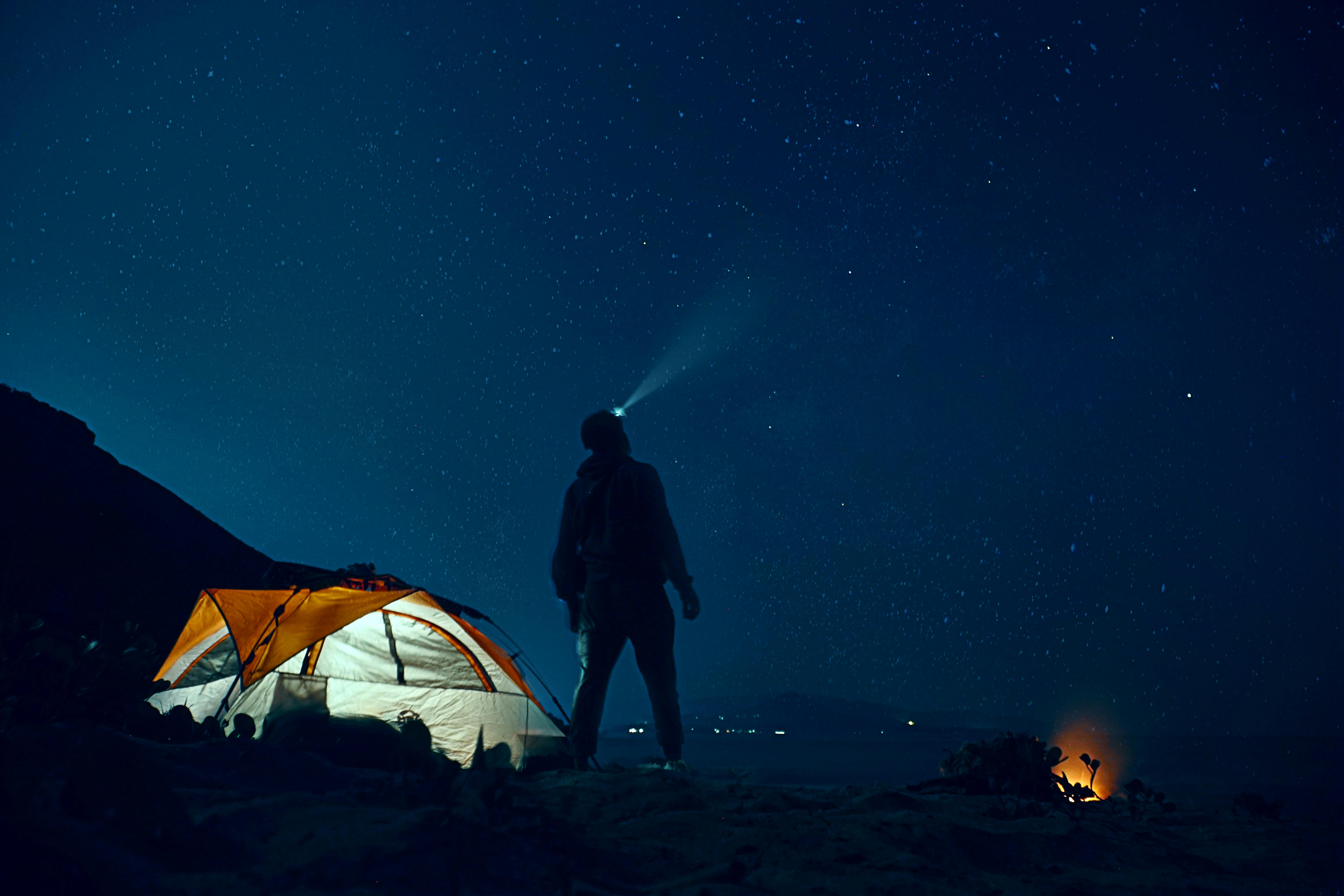 A lit up tent glows in the darkness while a figure with a headlamp stands next to it