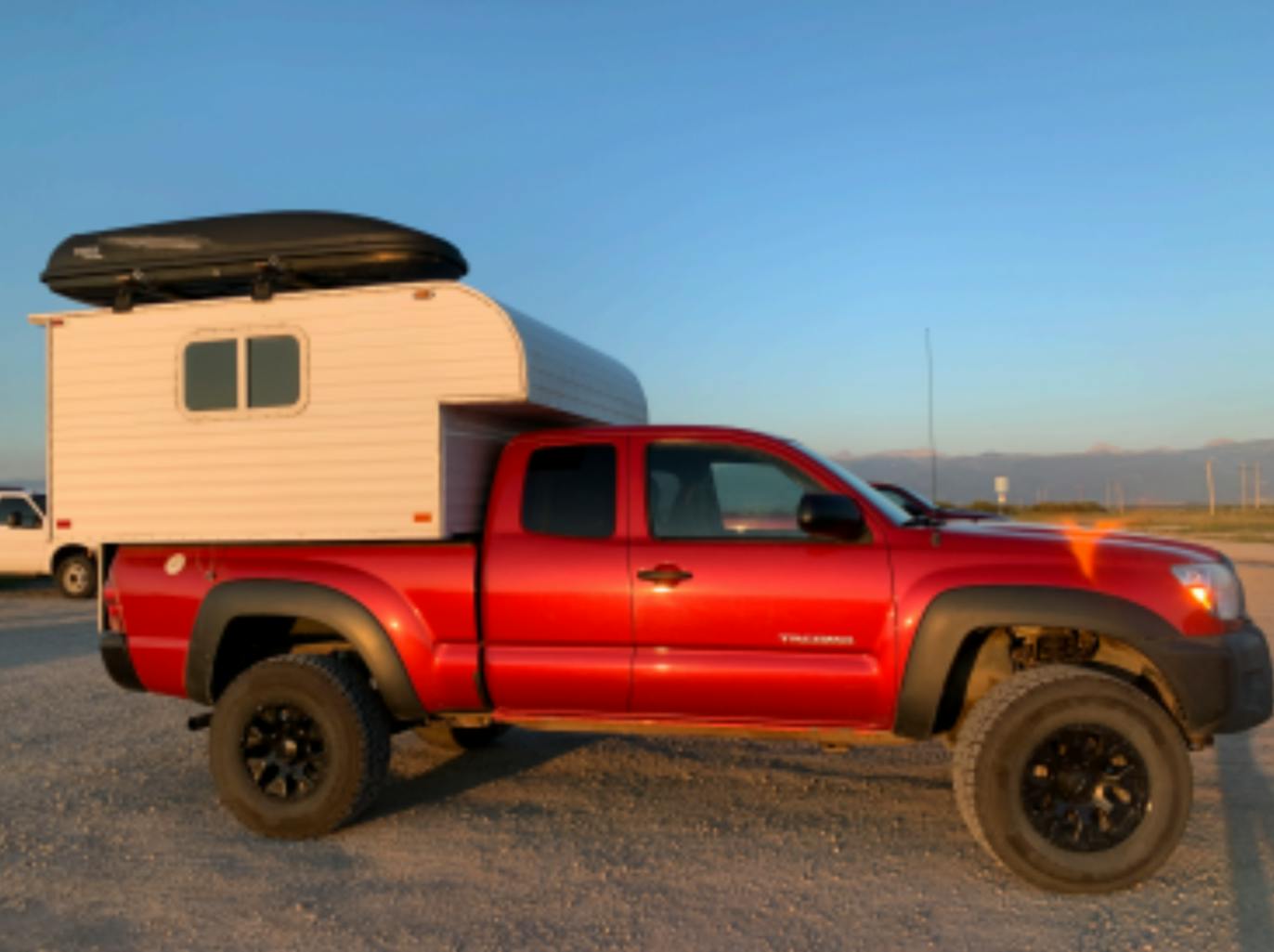 Red Toyota Tacoma truck with a white camper in the bed of the truck. The camper has a black storage box on top of it and the tires are black.