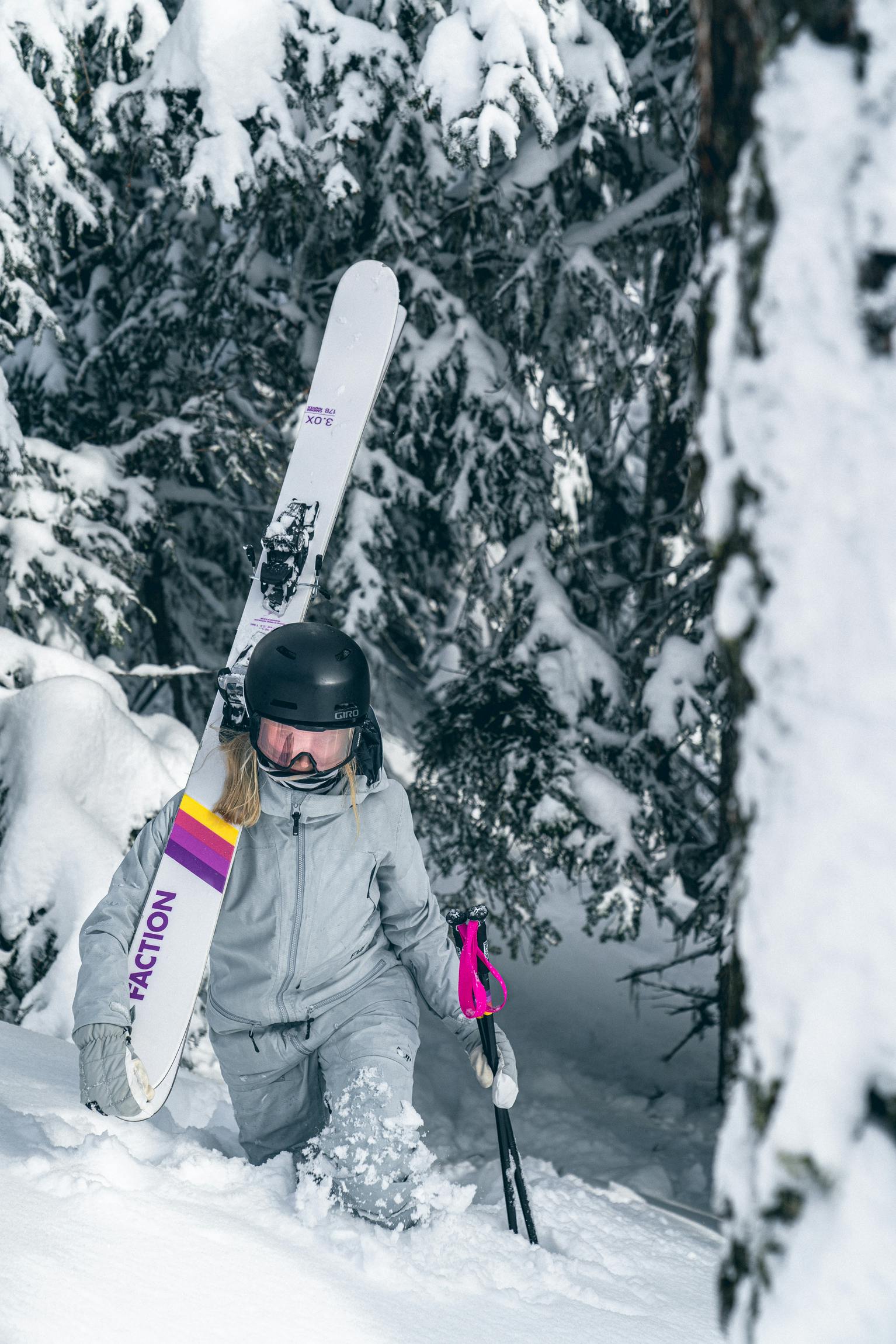 A woman carrying Faction skis and hiking up a slope in knee-deep snow