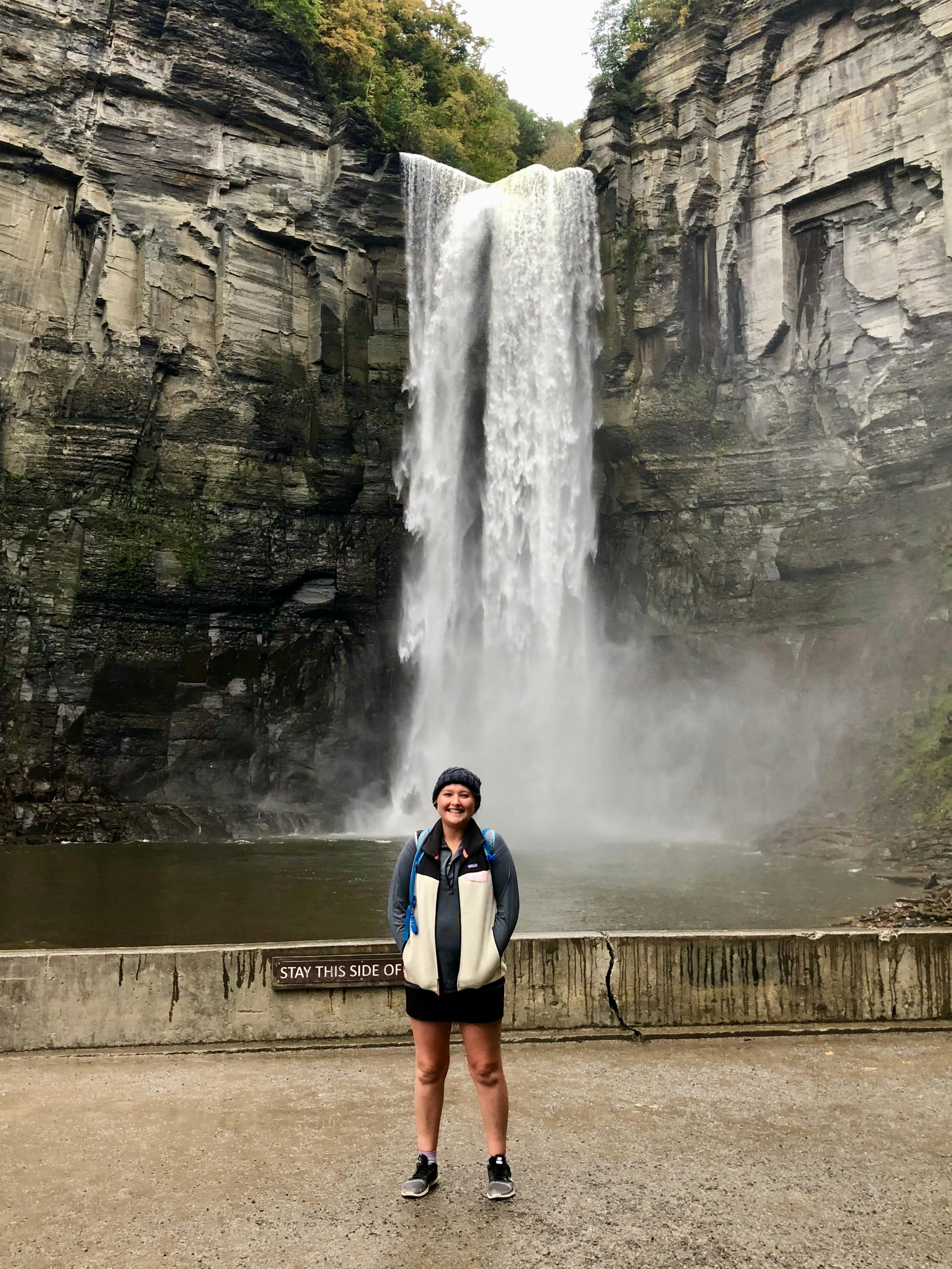 The author wears a hiking skirt, a fleece, and a beanie while standing on a platform in front of a waterfall.