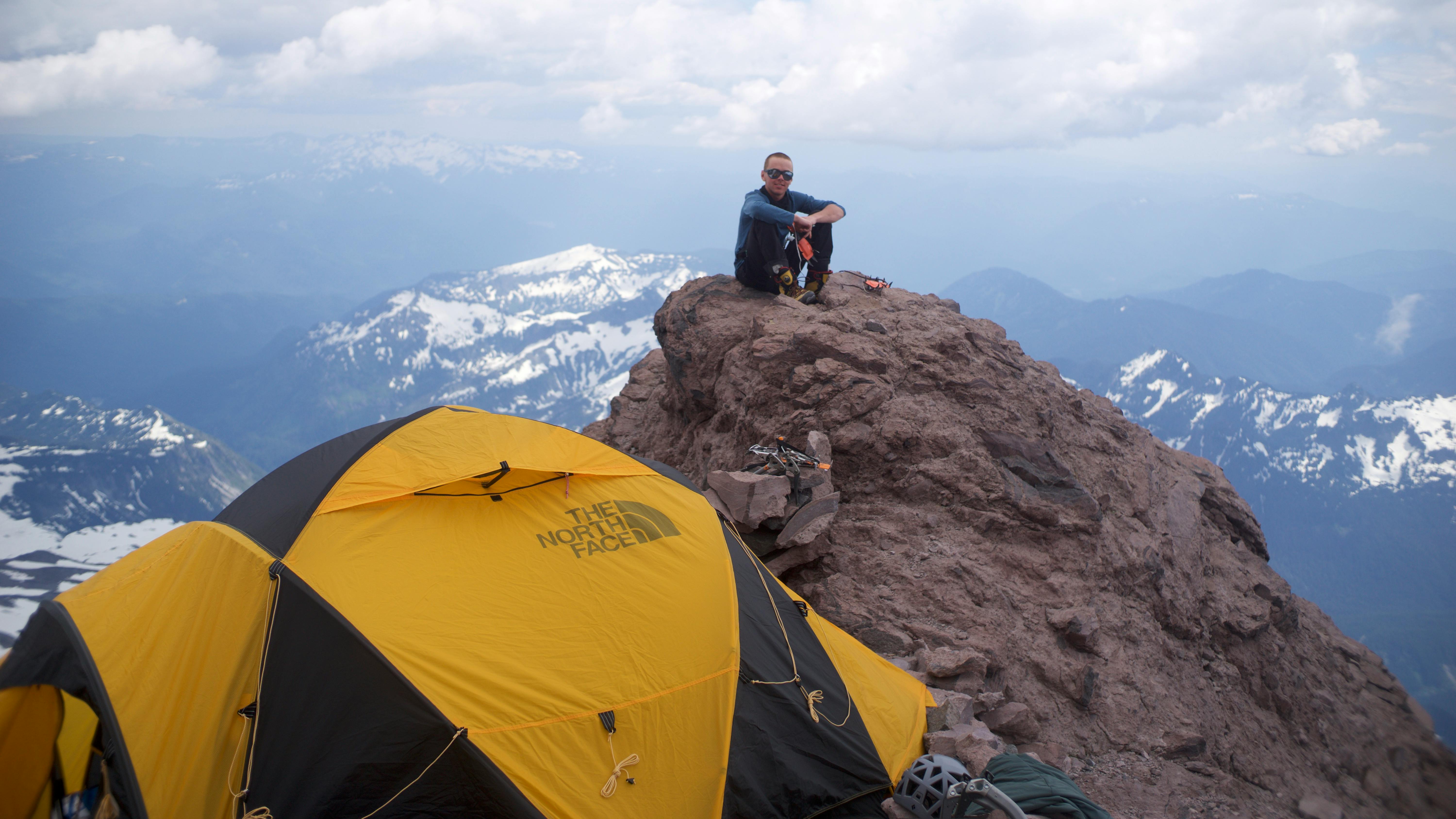 A man sits on a rock in front of snowy mountains with a yellow tent to his left