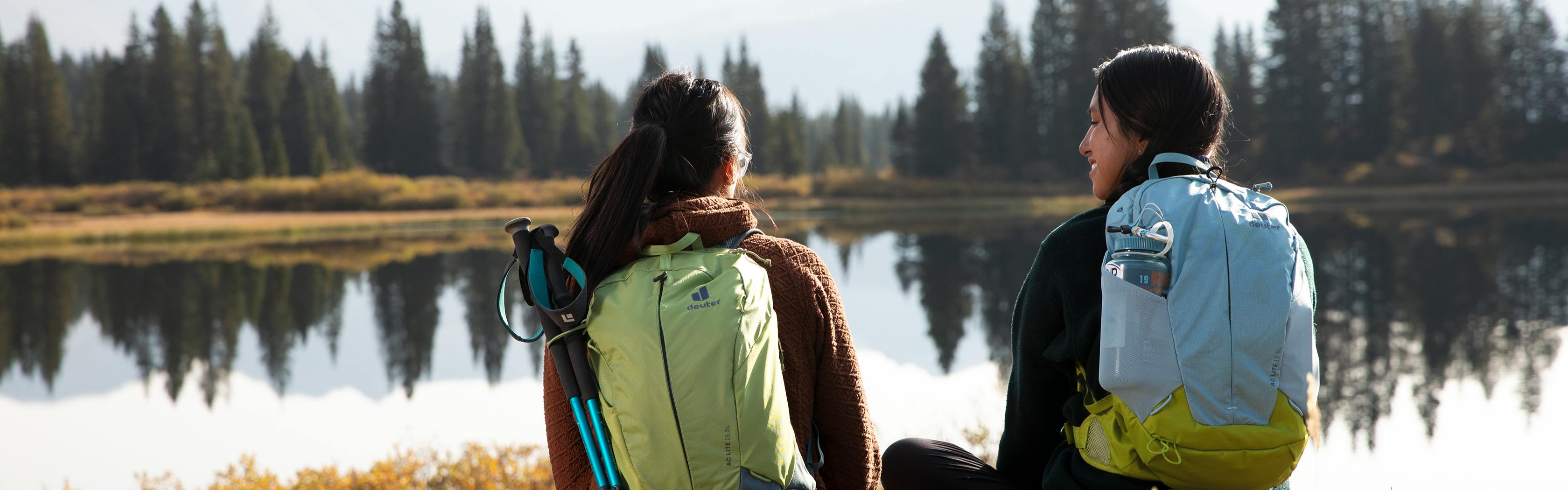 Two women wearing Deuter AC Lite backpacks sit in front of a lake.