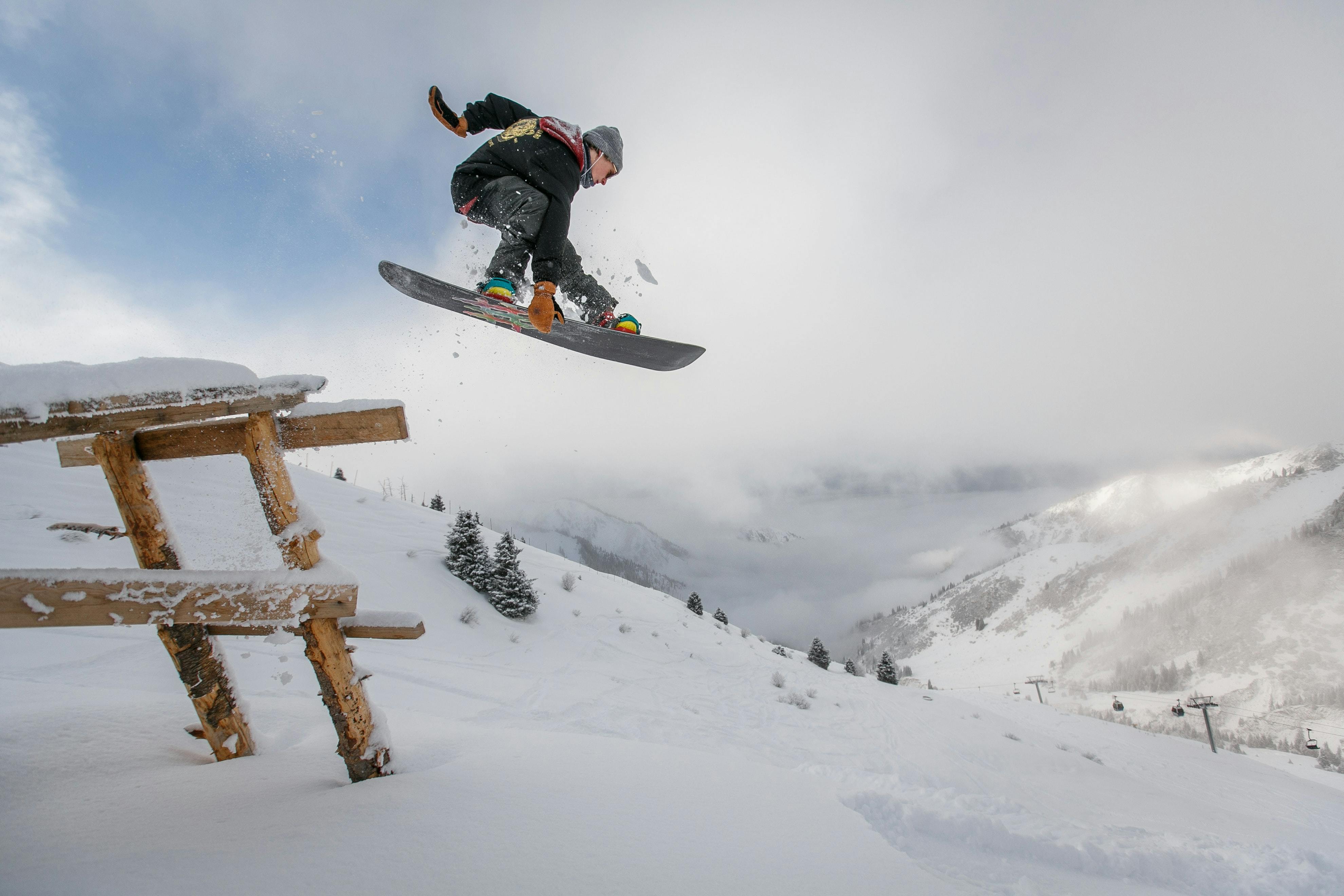 A snowboarder jumps over a wooden structure. He is really high in the air and grabbing his snowboard. There is a lot of snow on the ground and its cloudy outside. 