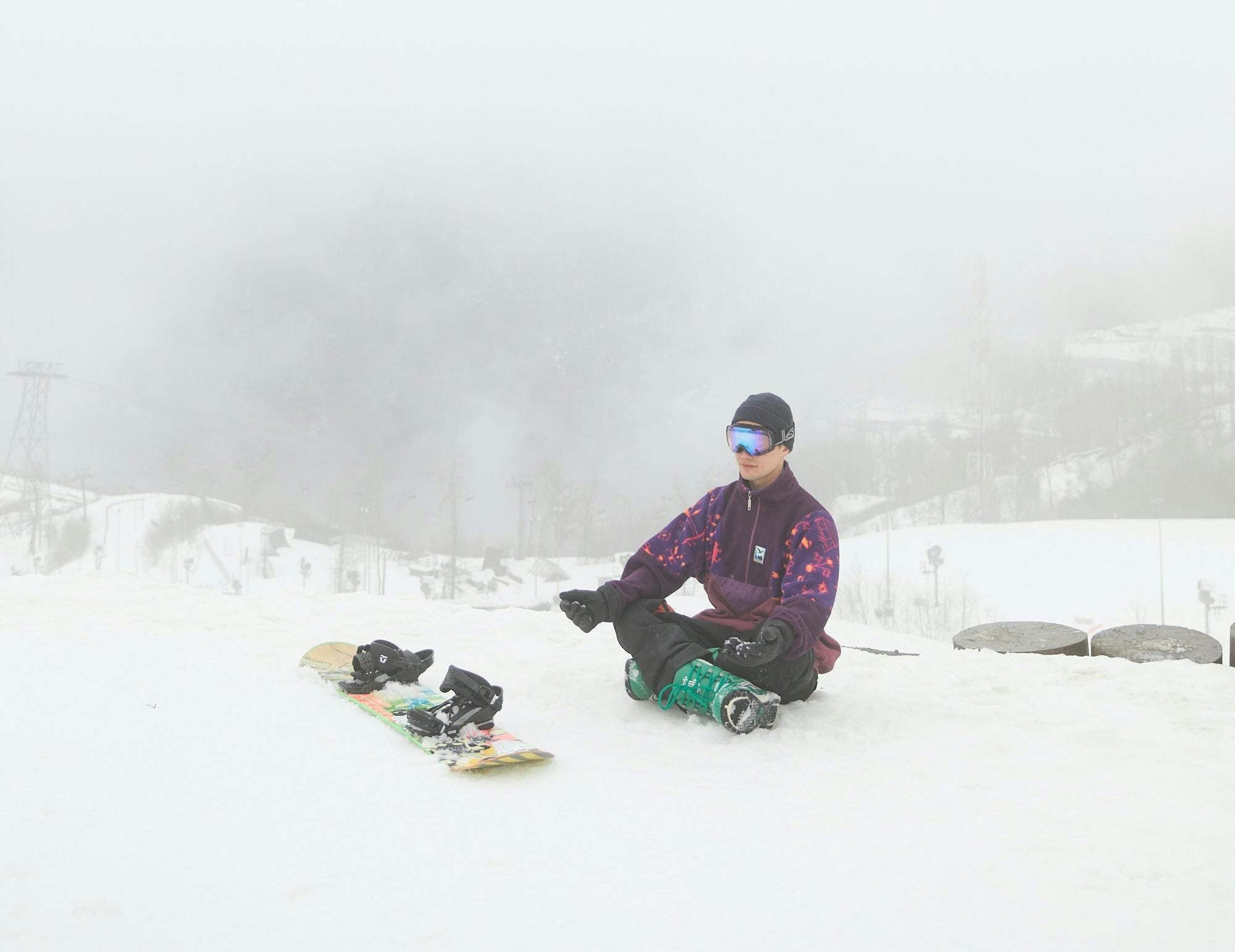 A man sits with his snowboard in front of him. It is gloomy outside and there is a lot of snow on the ground.