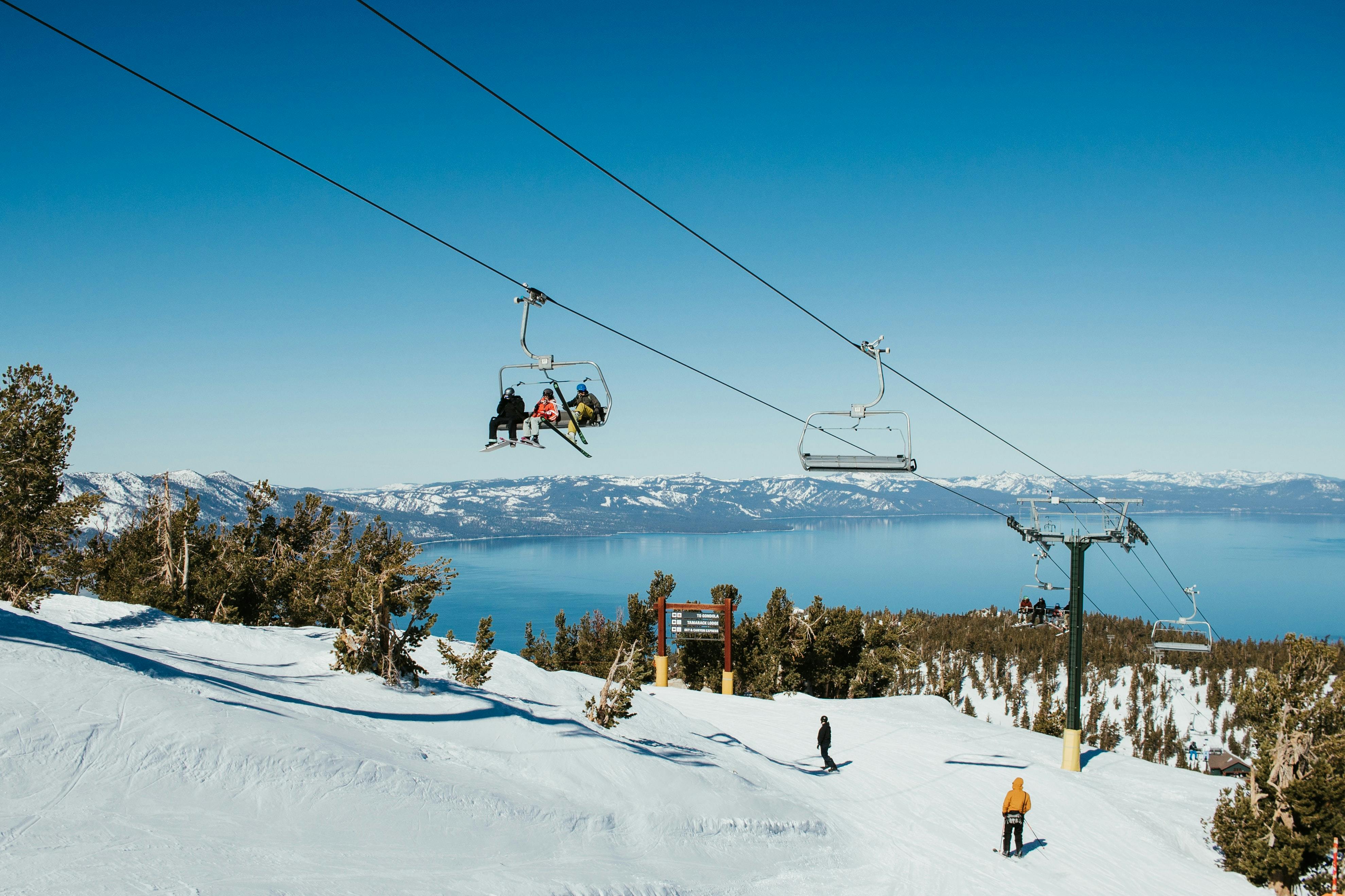View of a ski chairlift and ski run both with skiers on them. There is a a lake in the background. 