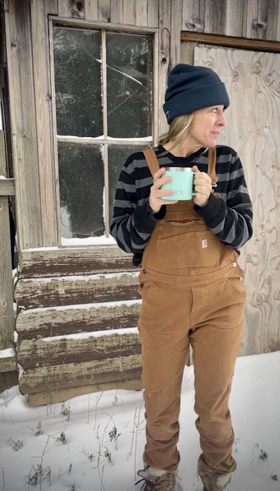 The author stands in the snow in Carhartt overalls and holds her mug. 