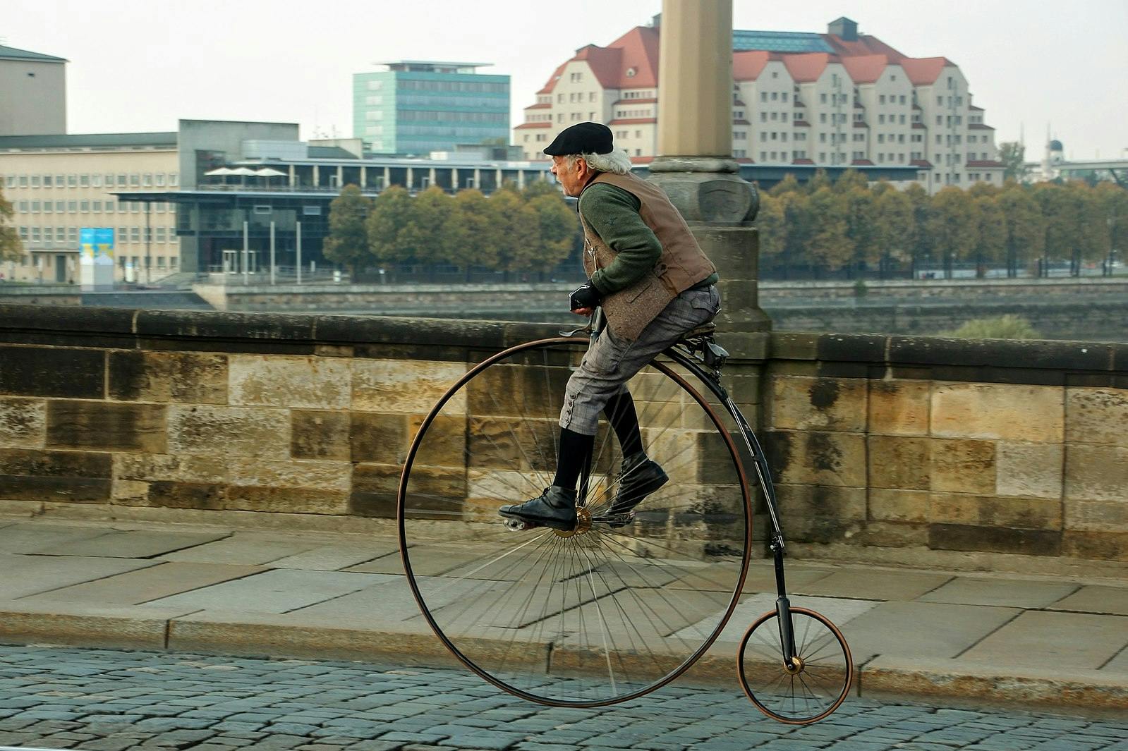 Old man crossing the Augustus bridge in Dresden, Germany on a penny-farthing bicycle. He is wearing a black hat and a brown vest.