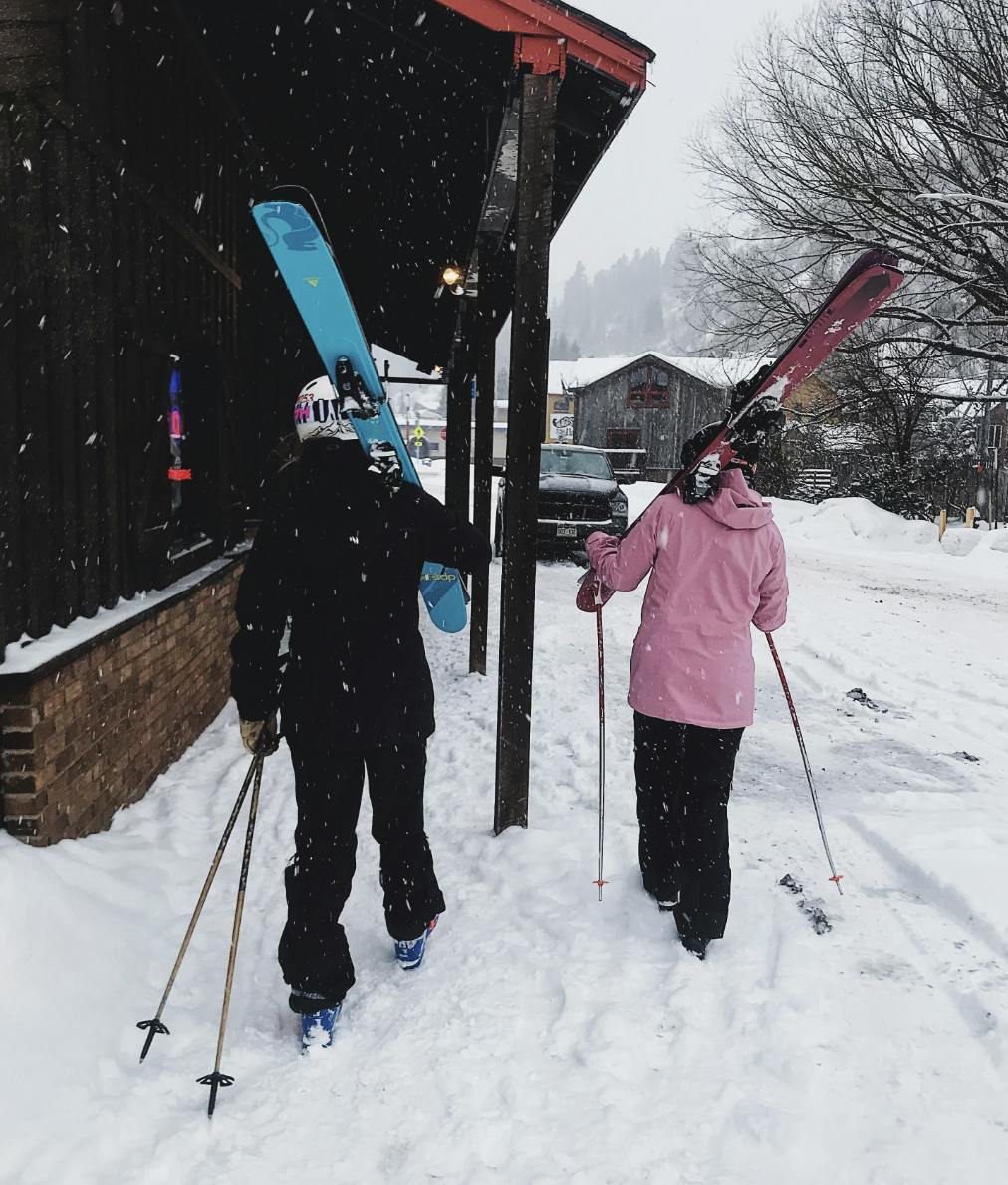 Two skiers walking in the snow. One is wearing the Flylow Women's Sphinx Bib Pants.