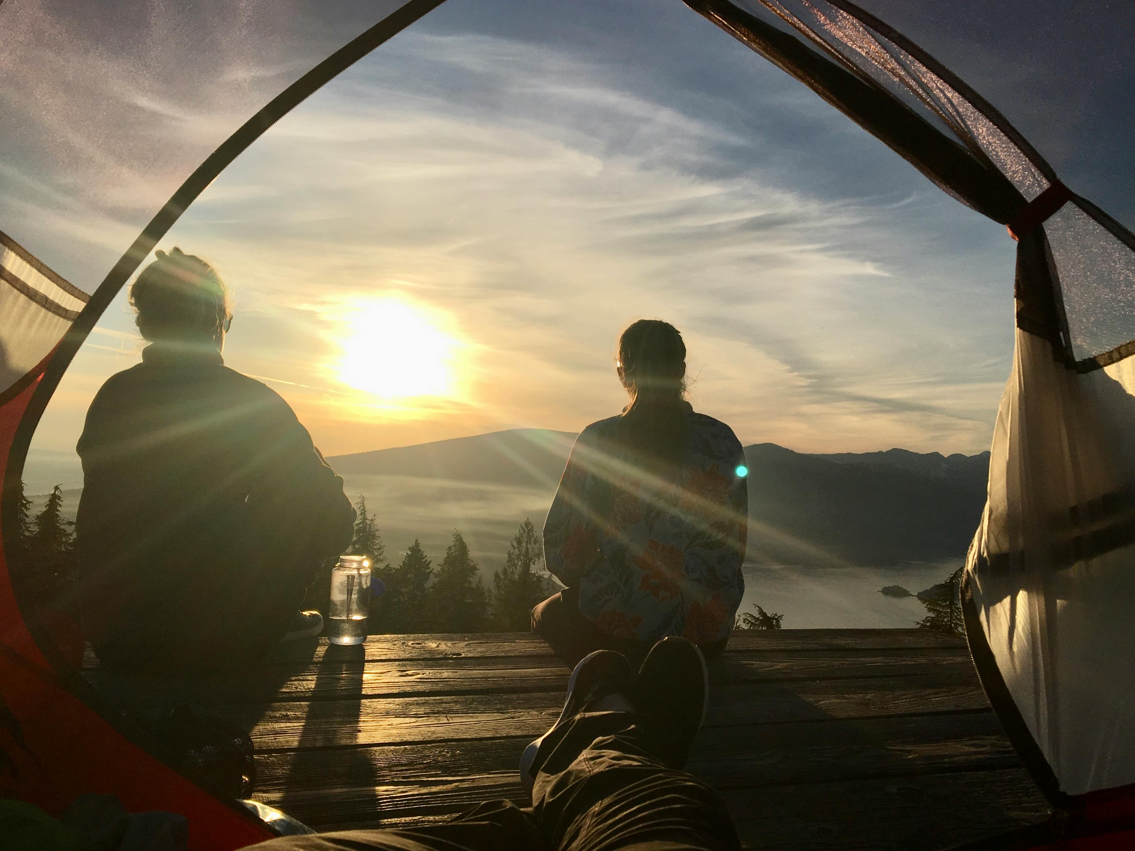Sitting in front of their tent, two hikers gaze out at a lake, mountains, and setting sun.