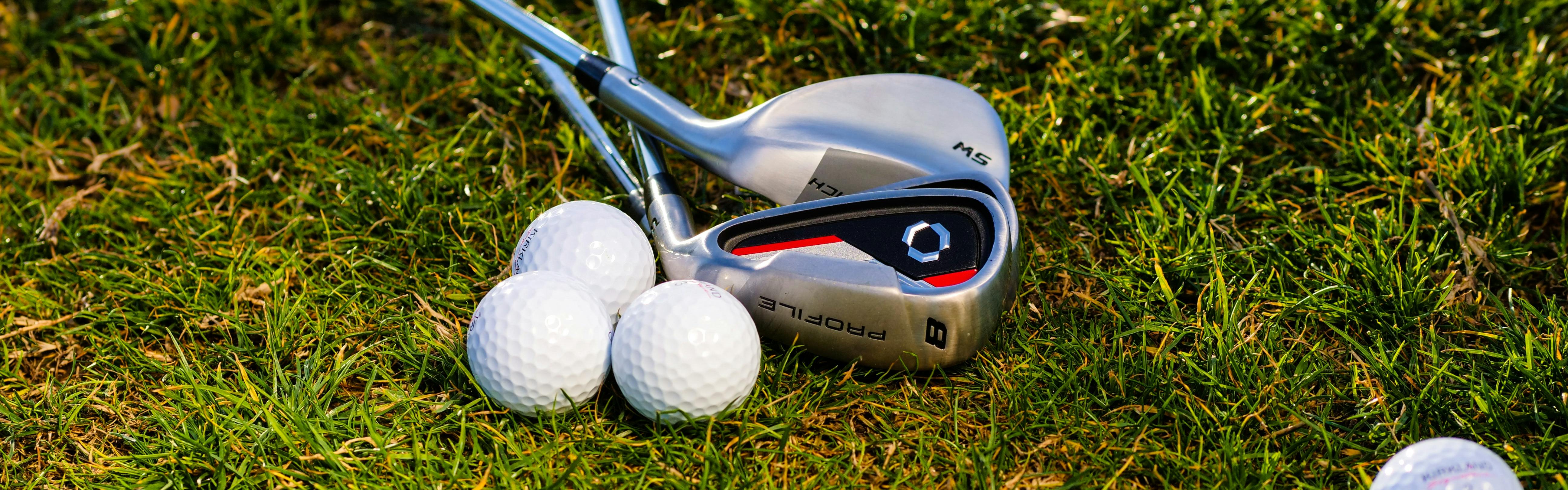 Two golf irons lying in the grass next to three golf balls. 
