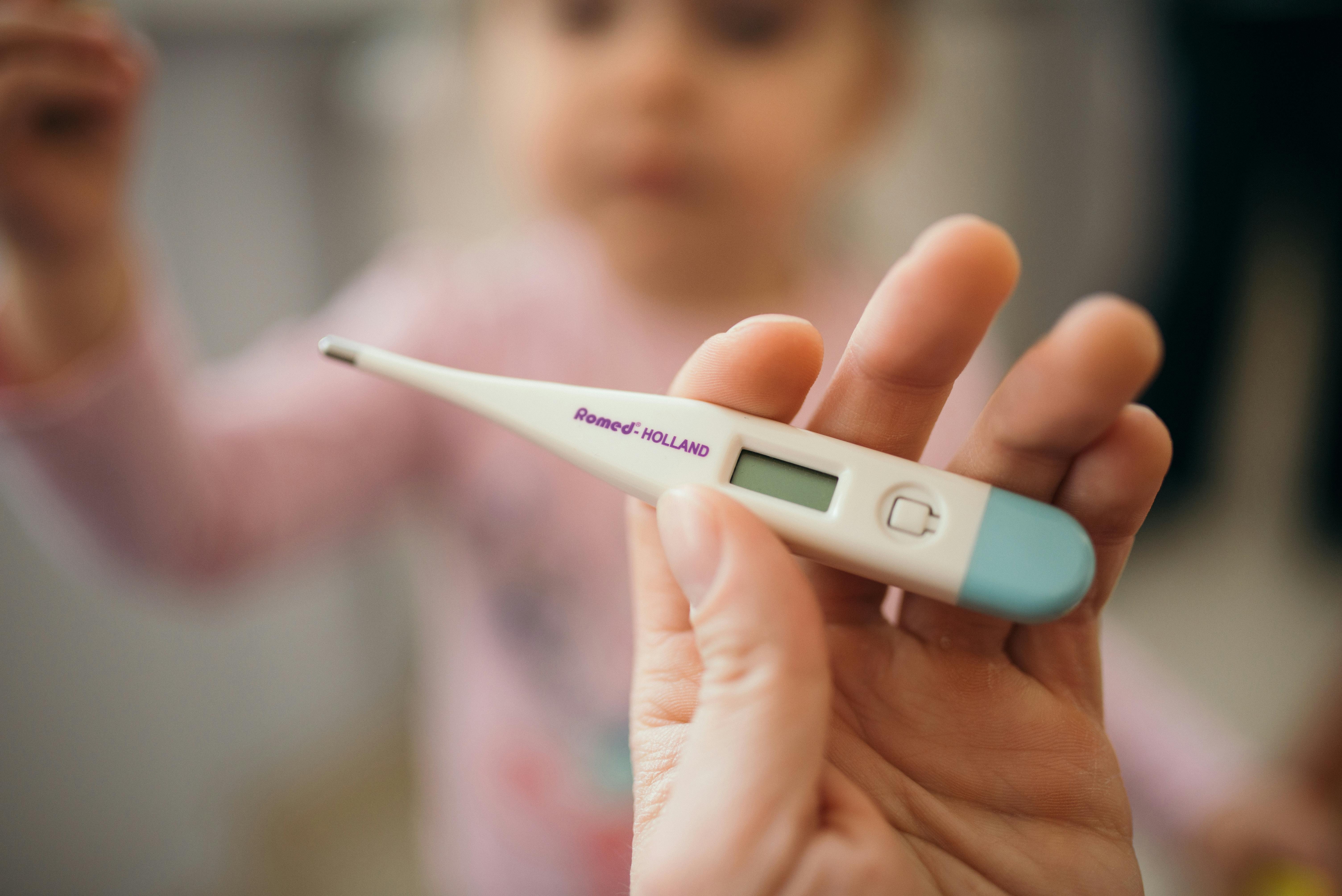 A hand holding a thermometer with a child in the background. 