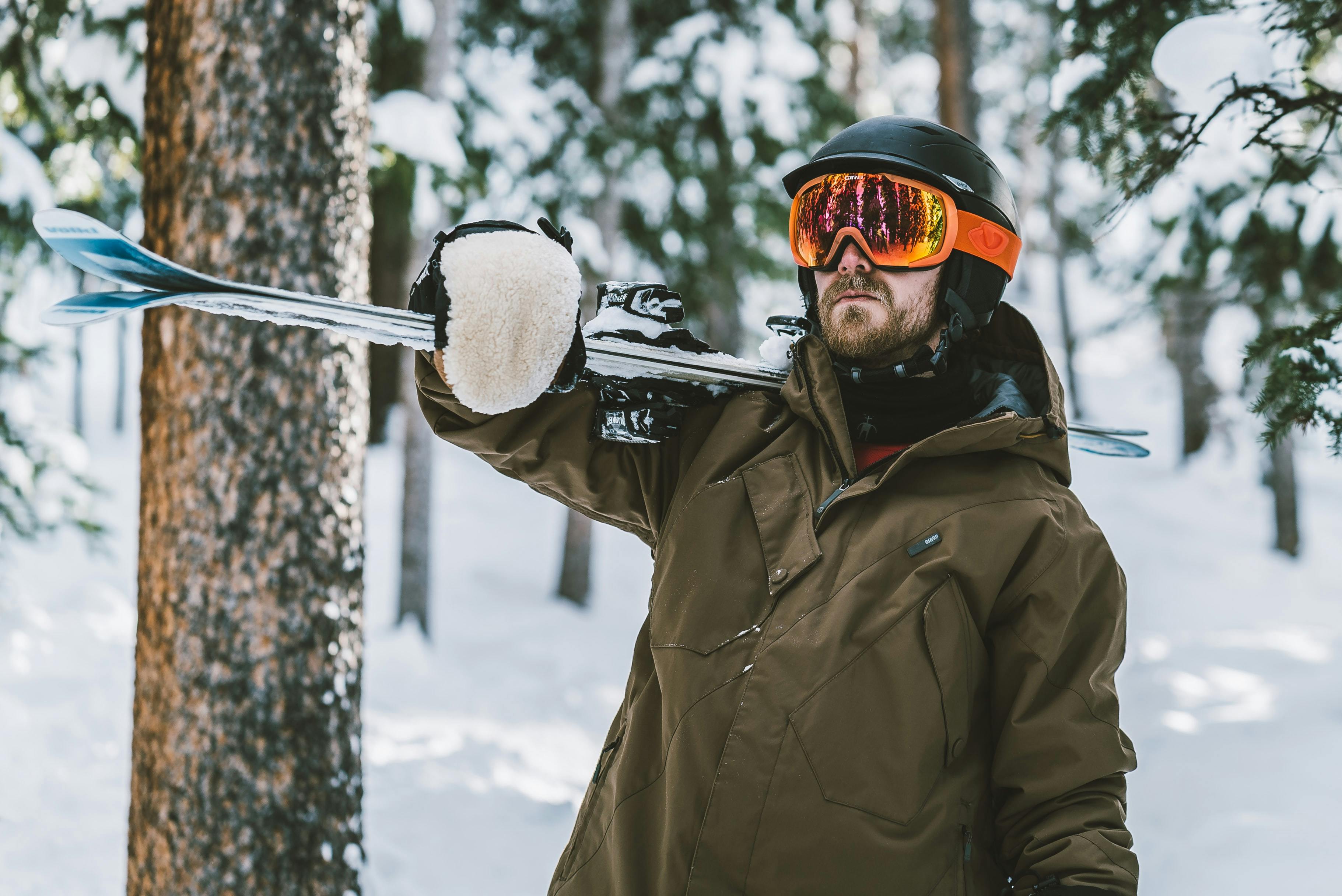A man holds skis over his shoulder. He is wearing a jacket, mittens, goggles, and a helmet.