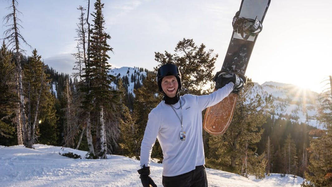 A man holding up a snowboard with snowy mountains behind him, 