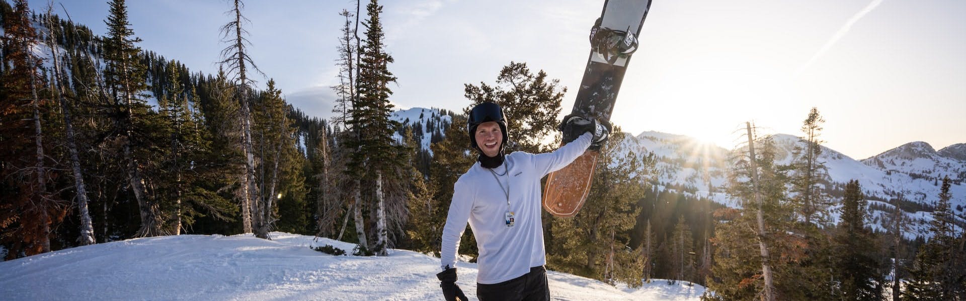 A man holding up a snowboard with snowy mountains behind him, 