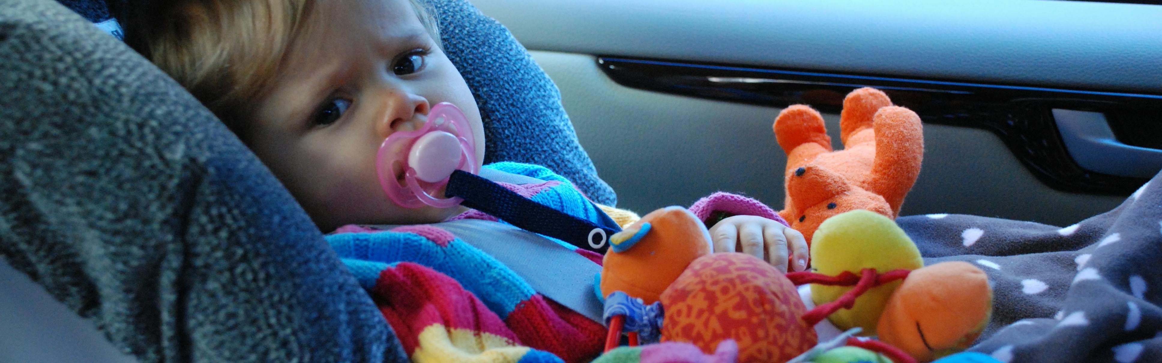 A baby in a car seat looks at the camera. The baby is holding toys and sucking on a pacifier. 