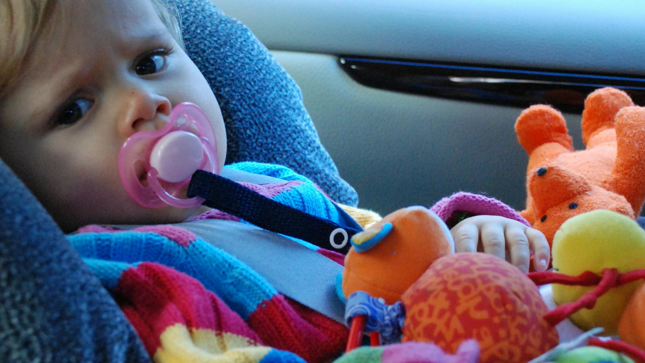 A baby in a car seat looks at the camera. The baby is holding toys and sucking on a pacifier. 