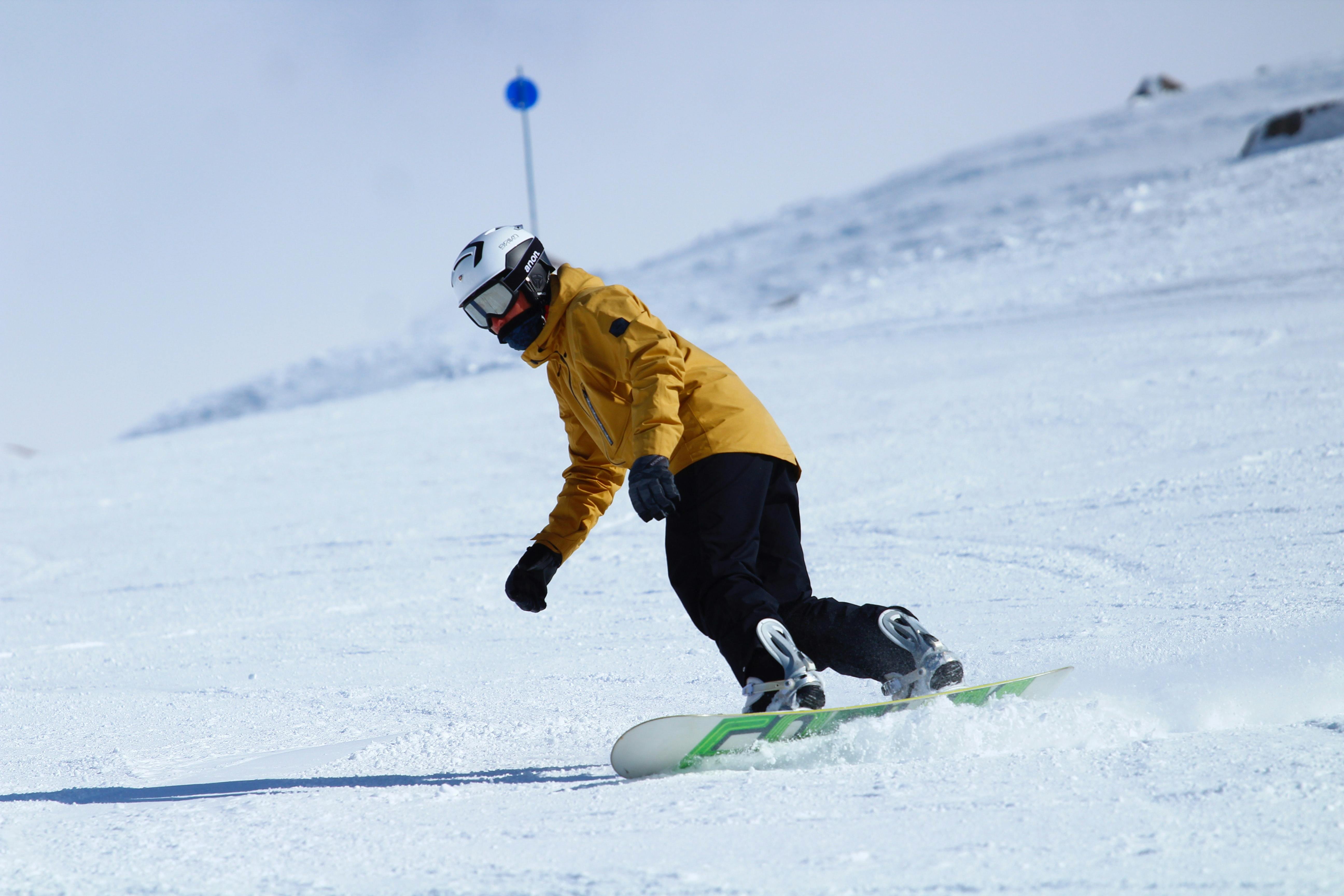 Someone rides a snowboard in a helmet, goggles, mittens, a yellow jacket, and black snow pants. 