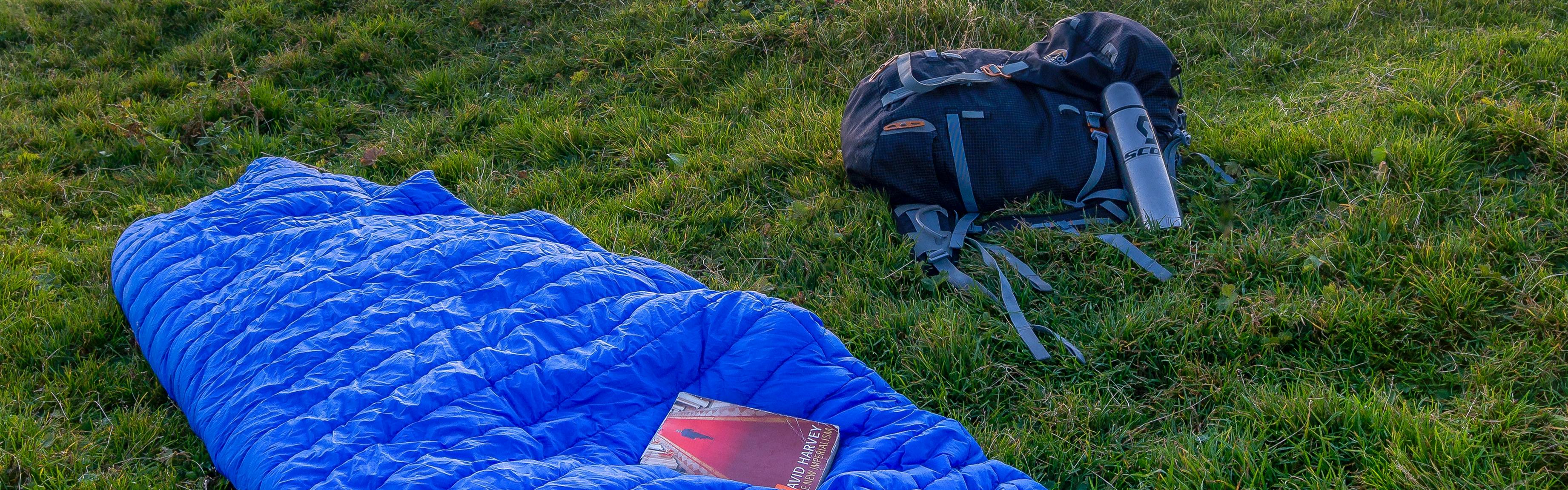 A navy backpack lays next to a royal blue sleeping bag on grassy ground. A book is sitting on top of the sleeping bag. 