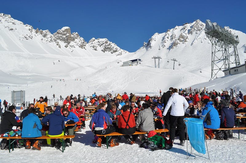 Several skiers sit at tables in a base area at a ski resort. 