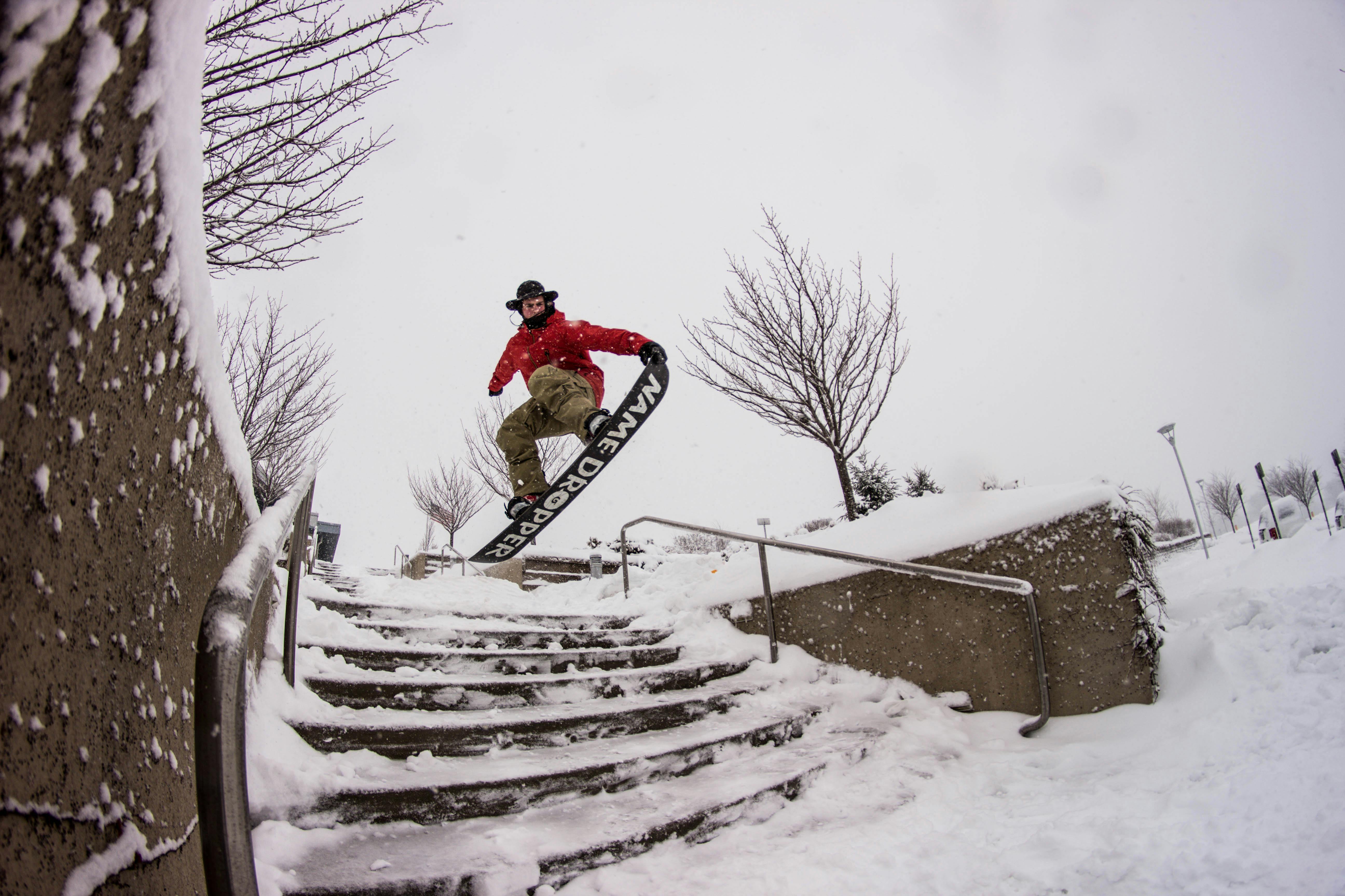 A snowboarder in a red jacket executes a jump down snowy stairs