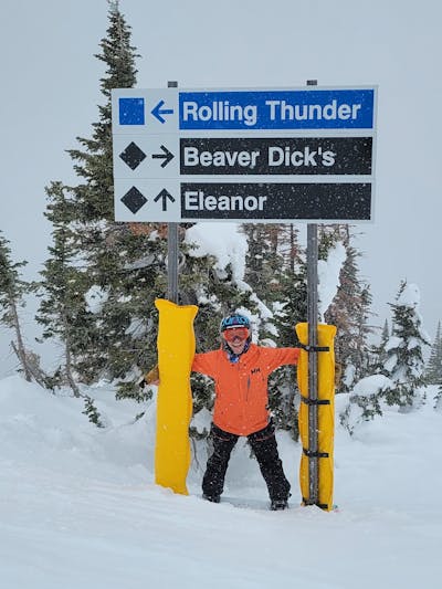 A snowboarder standing under a chairlift sign. 