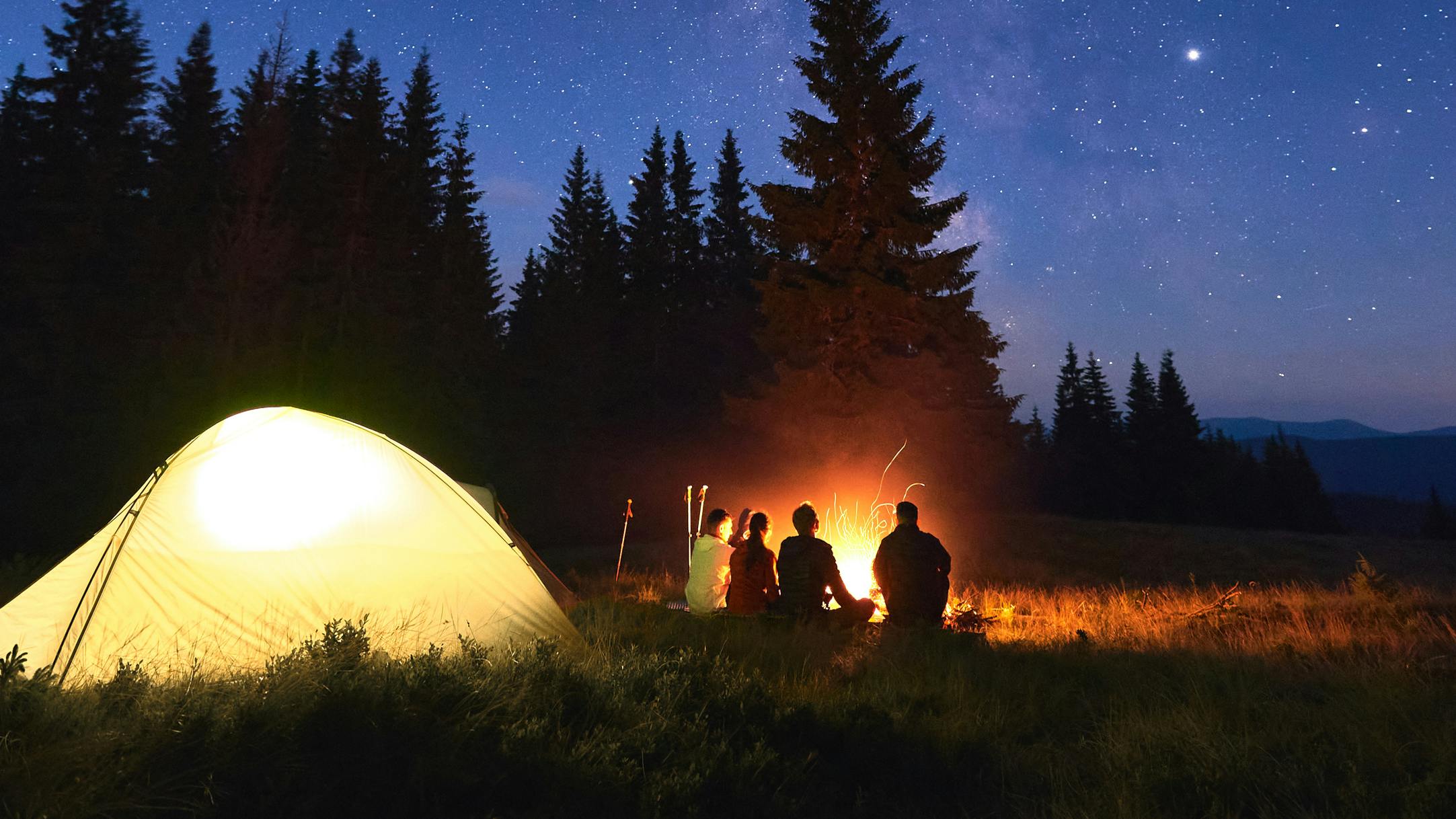 A group of people gather around a campfire at night while a tent glows behind them