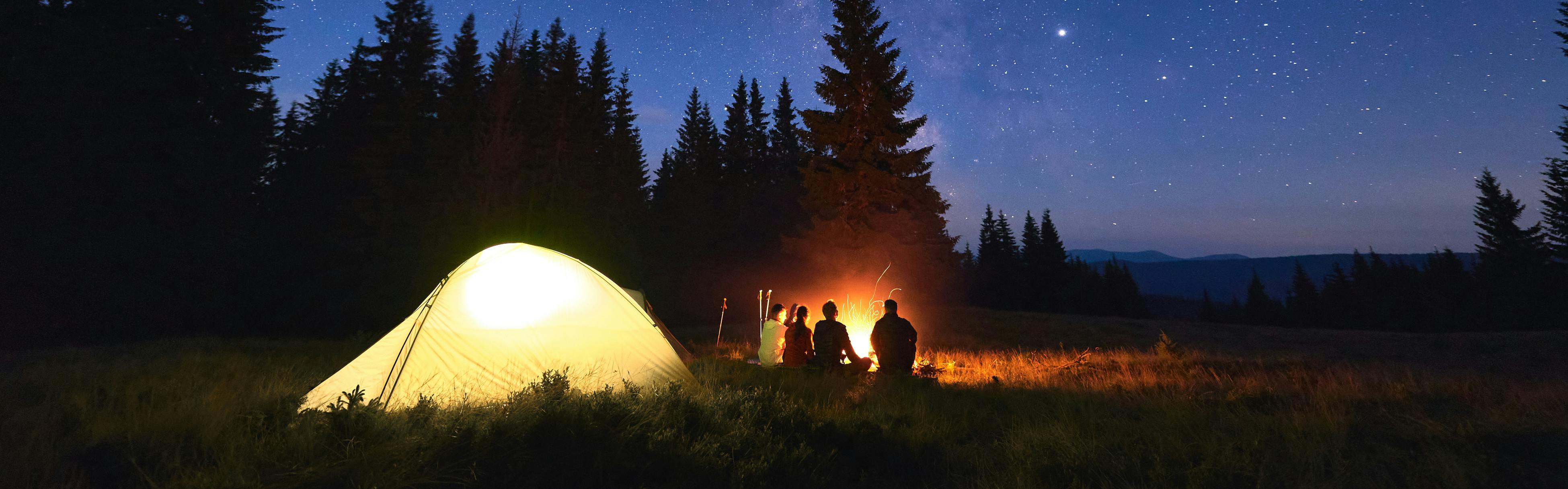 A group of people gather around a campfire at night while a tent glows behind them