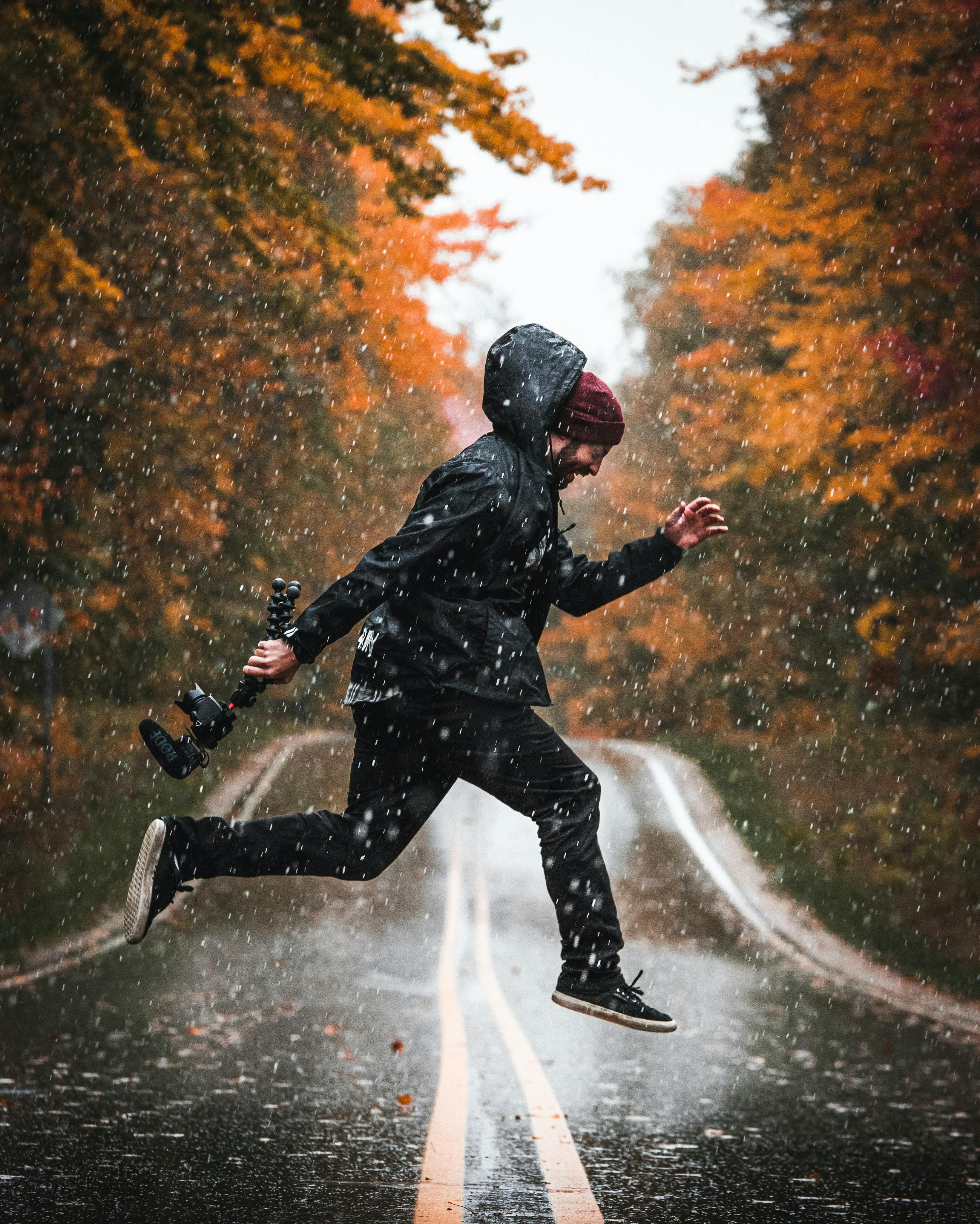 A man in a black hooded jacket jumping in the road in the rain with autumn foliage around him